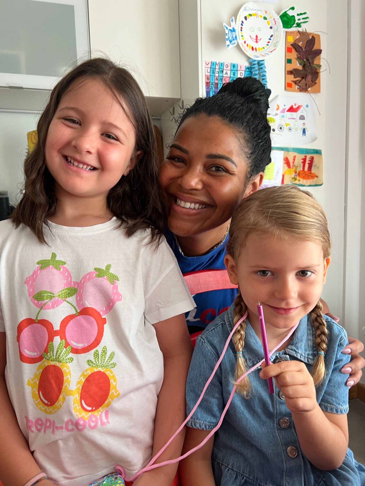 Two children and a woman smiling, with colorful artwork on the wall behind them, in a classroom setting.