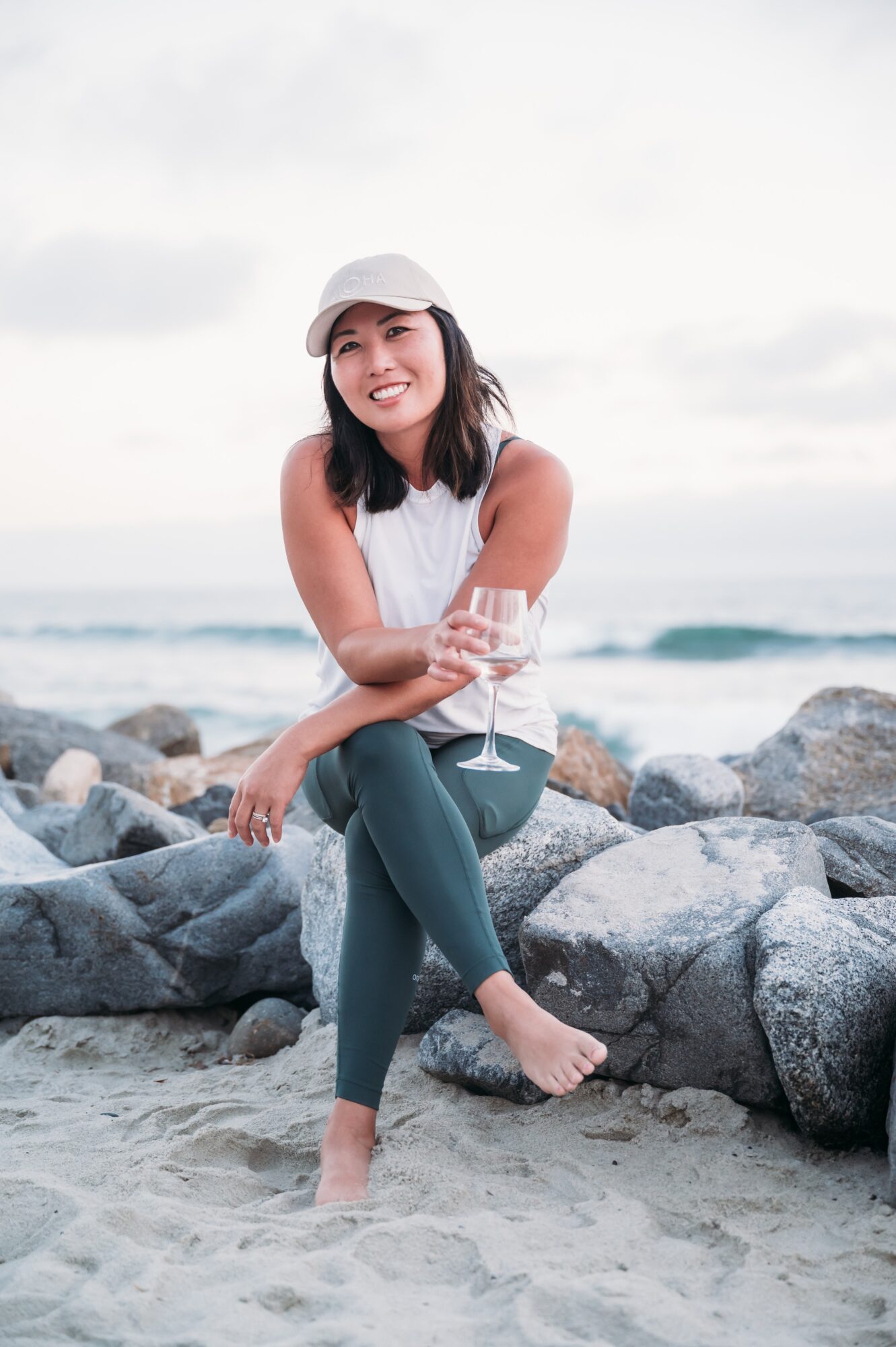 Woman sitting on rocks at the beach, smiling, holding a glass of wine, wearing a white hat and sleeveless top.