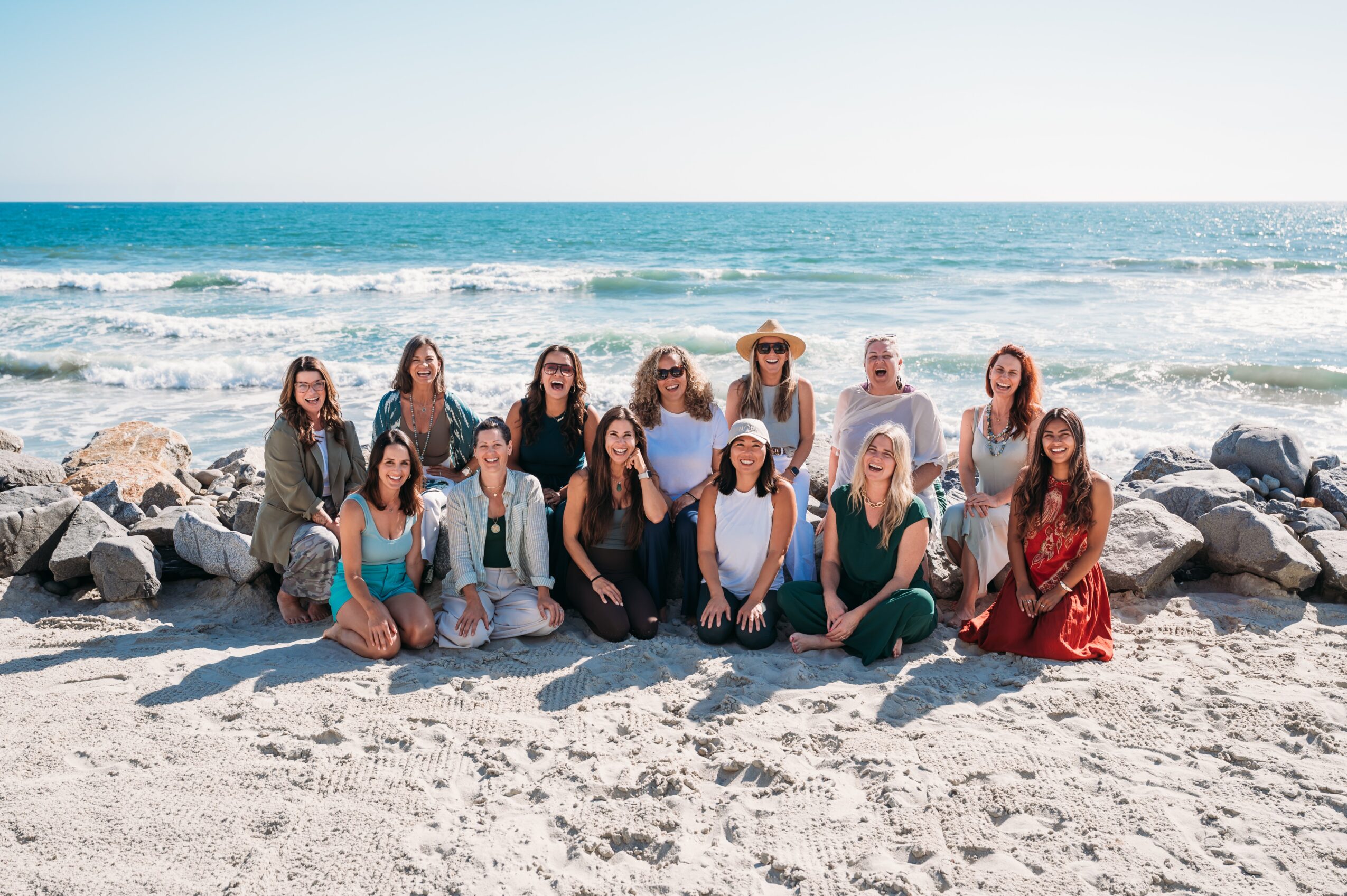 Group of fifteen women sitting and standing on a sandy beach with rocks and ocean behind them, smiling at the camera.