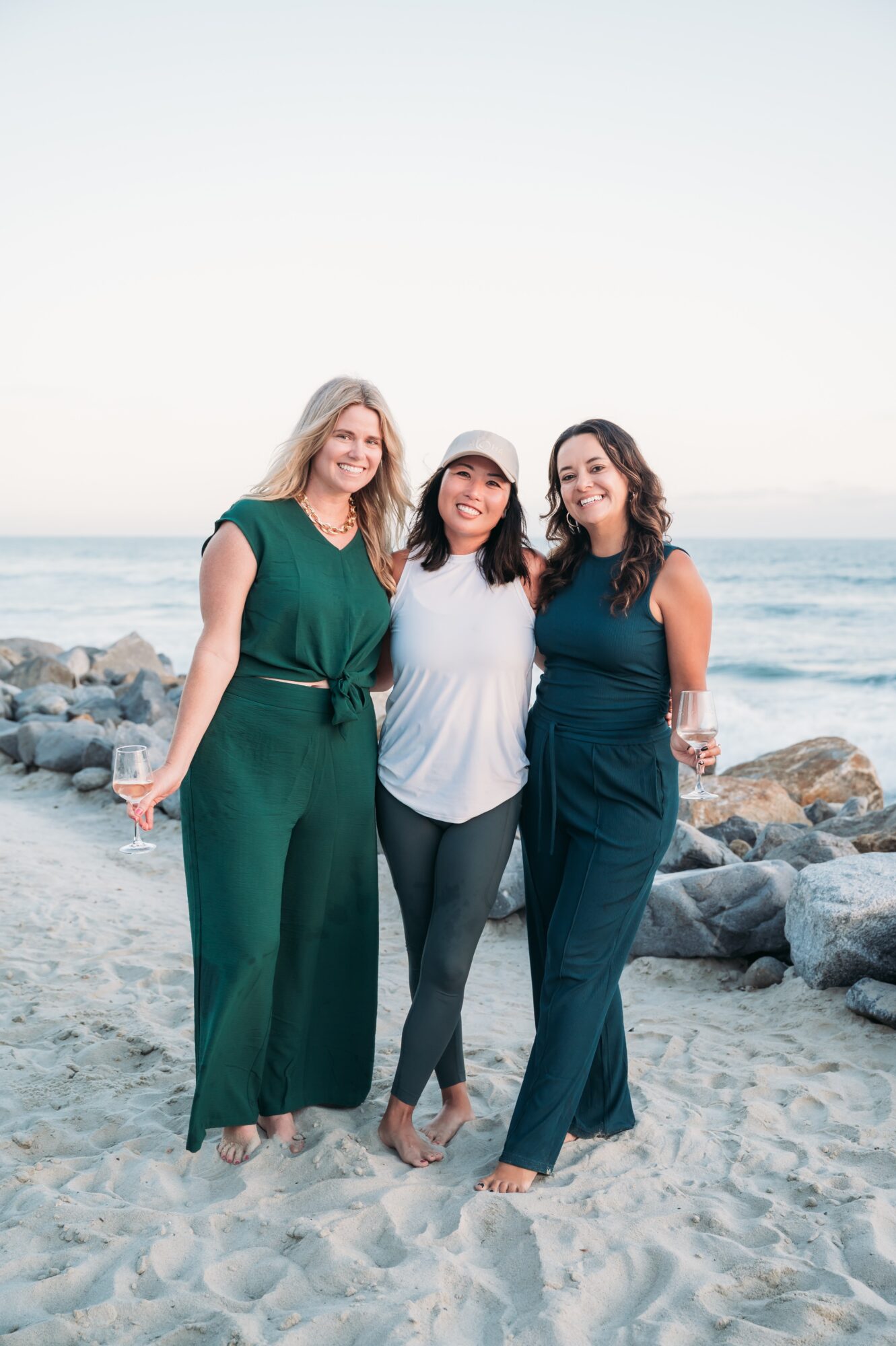 Three women standing on a beach with rocks and ocean in background, smiling, with arms around each other, wearing casual clothing.