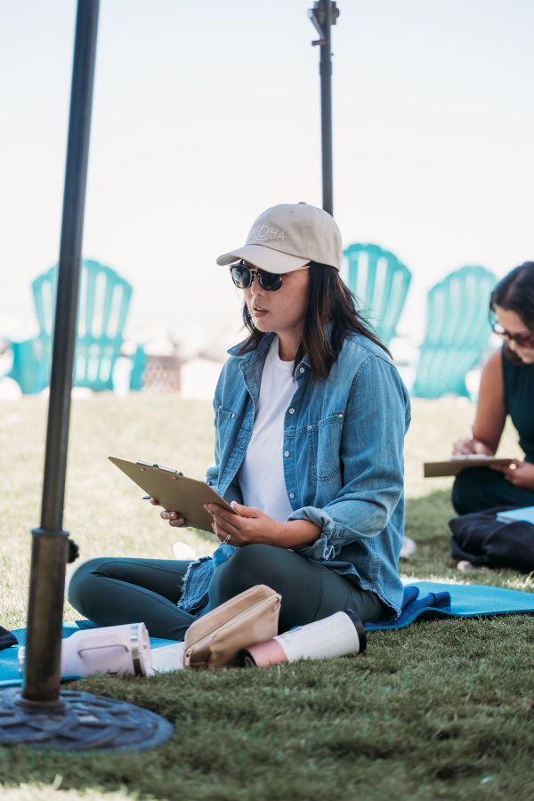 Woman sitting on grass reading a book outdoors, wearing sunglasses, a hat, and a denim jacket.