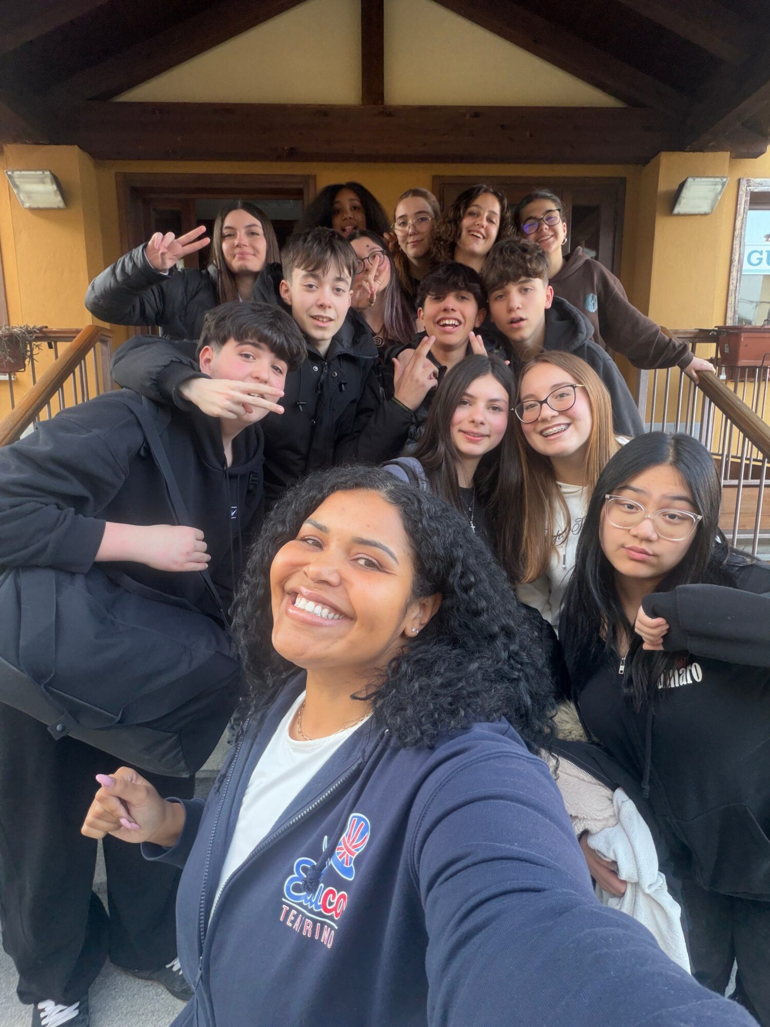 Group of young people smiling and posing for a photo indoors, some making peace signs, in a wooden structure.