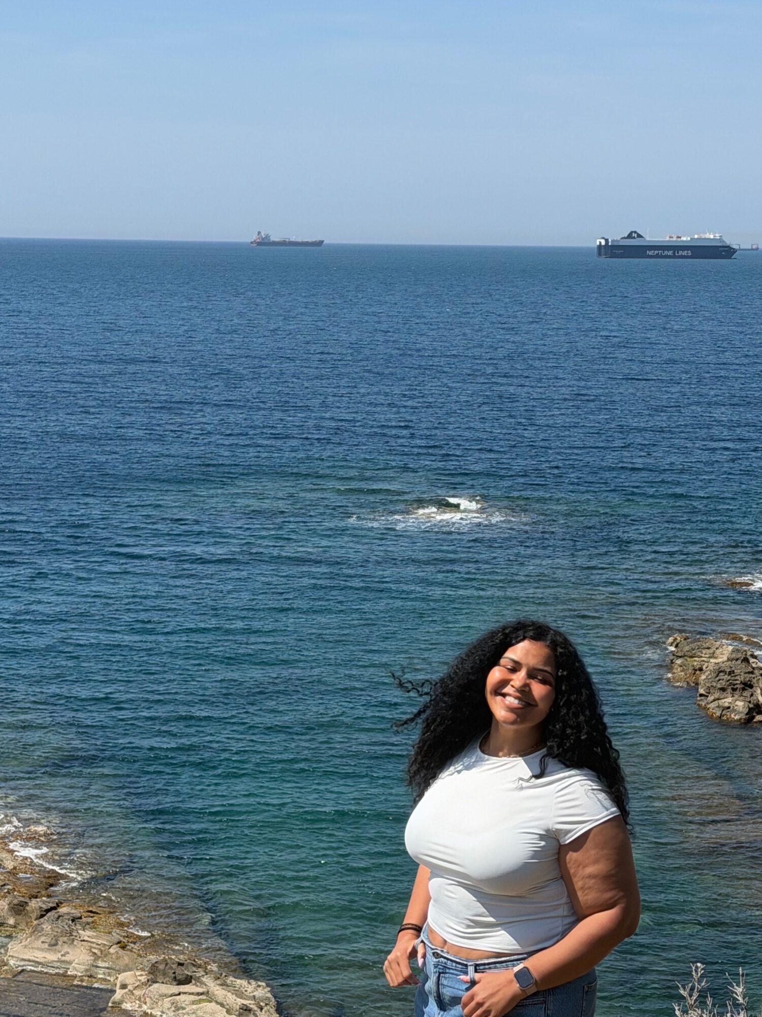 Woman standing on rocks near water, with a boat in the distance, under a clear sky.