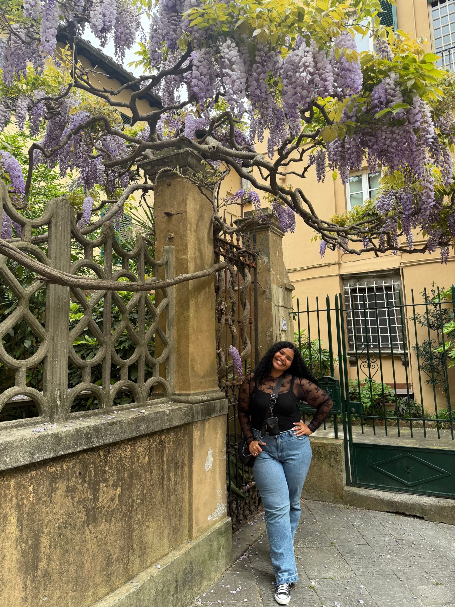 Woman standing on sidewalk near a tree with purple flowers, in front of a building with green door and window bars.