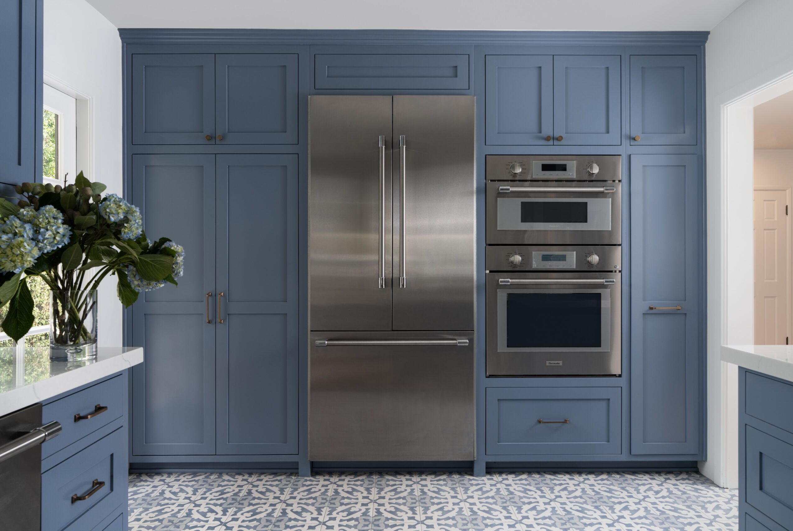 Kitchen with blue cabinets, stainless steel refrigerator, oven, and microwave, and a window with flowers on the left.