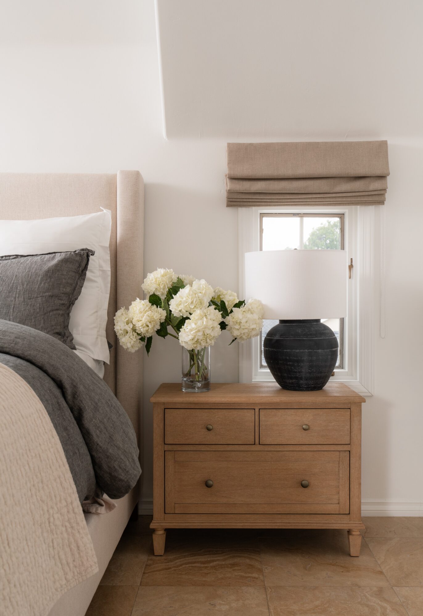 Nightstand with a vase of white flowers, a lamp, and a window with a beige Roman shade, next to a bed with gray bedding.