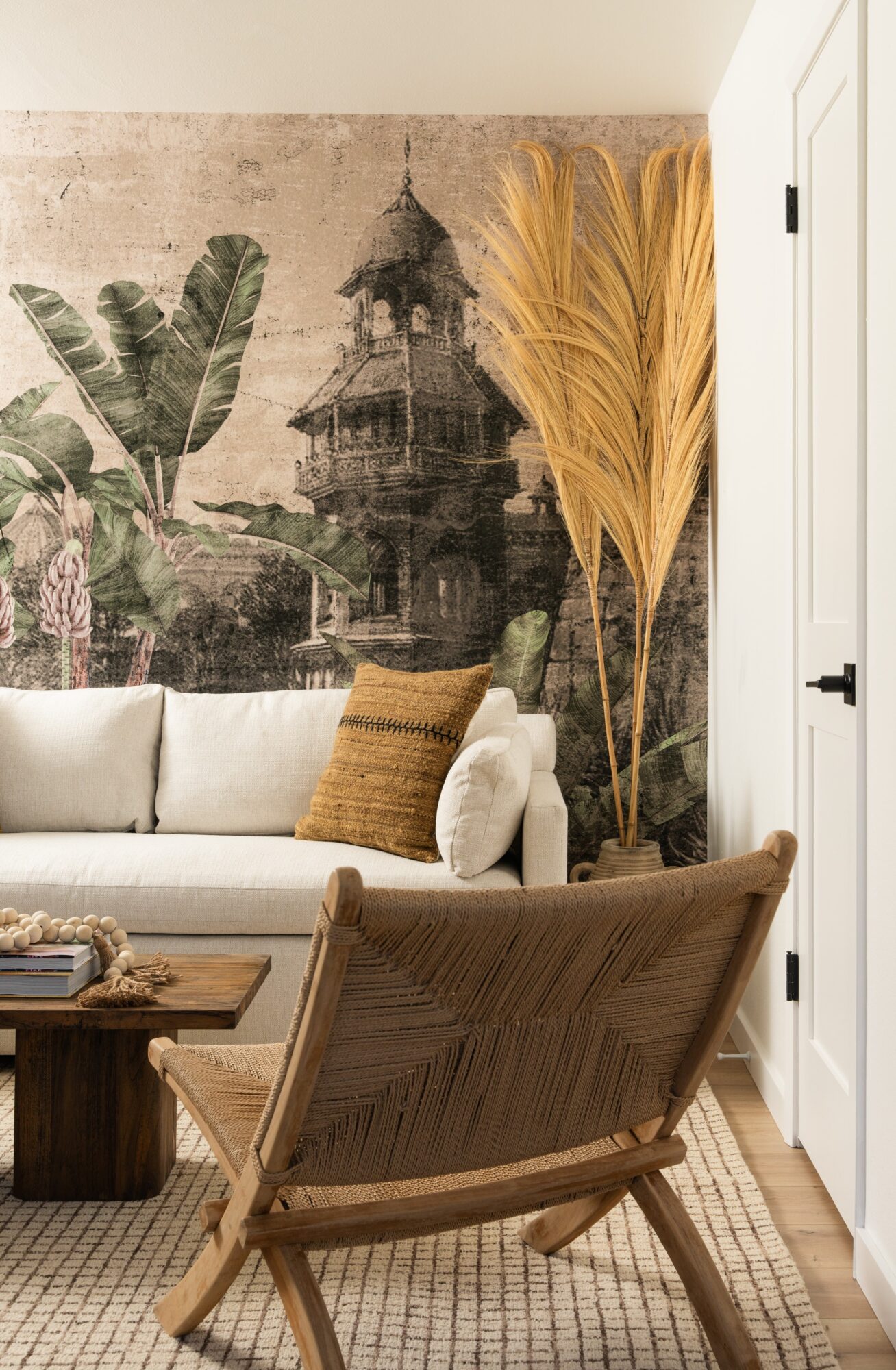 Living room with a white sofa, a brown pillow, a wooden chair, large dried plants, and a wall mural of a building and trees.