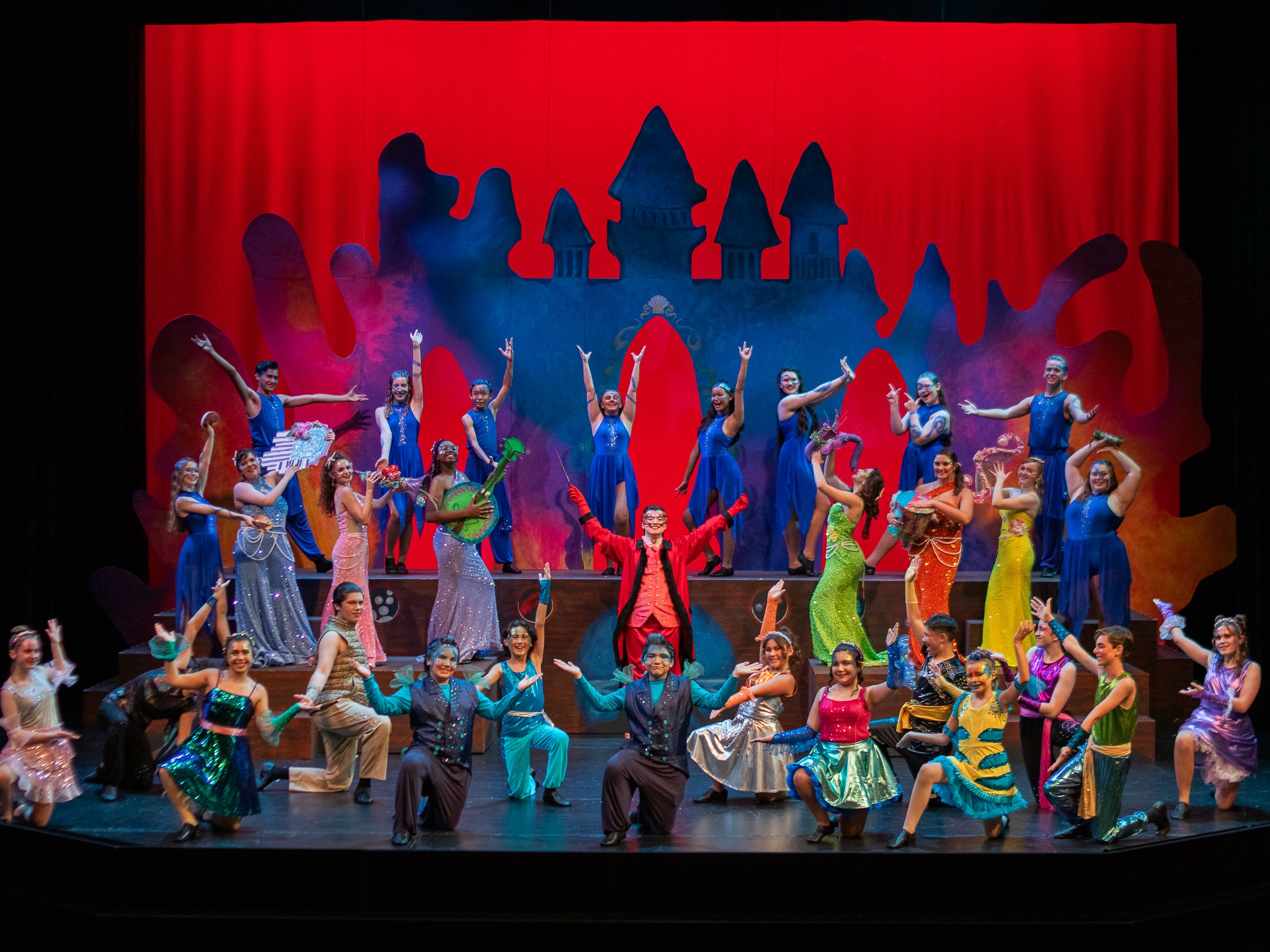 Group of performers on stage with colorful costumes, dancing in front of a red curtain and castle silhouette backdrop.