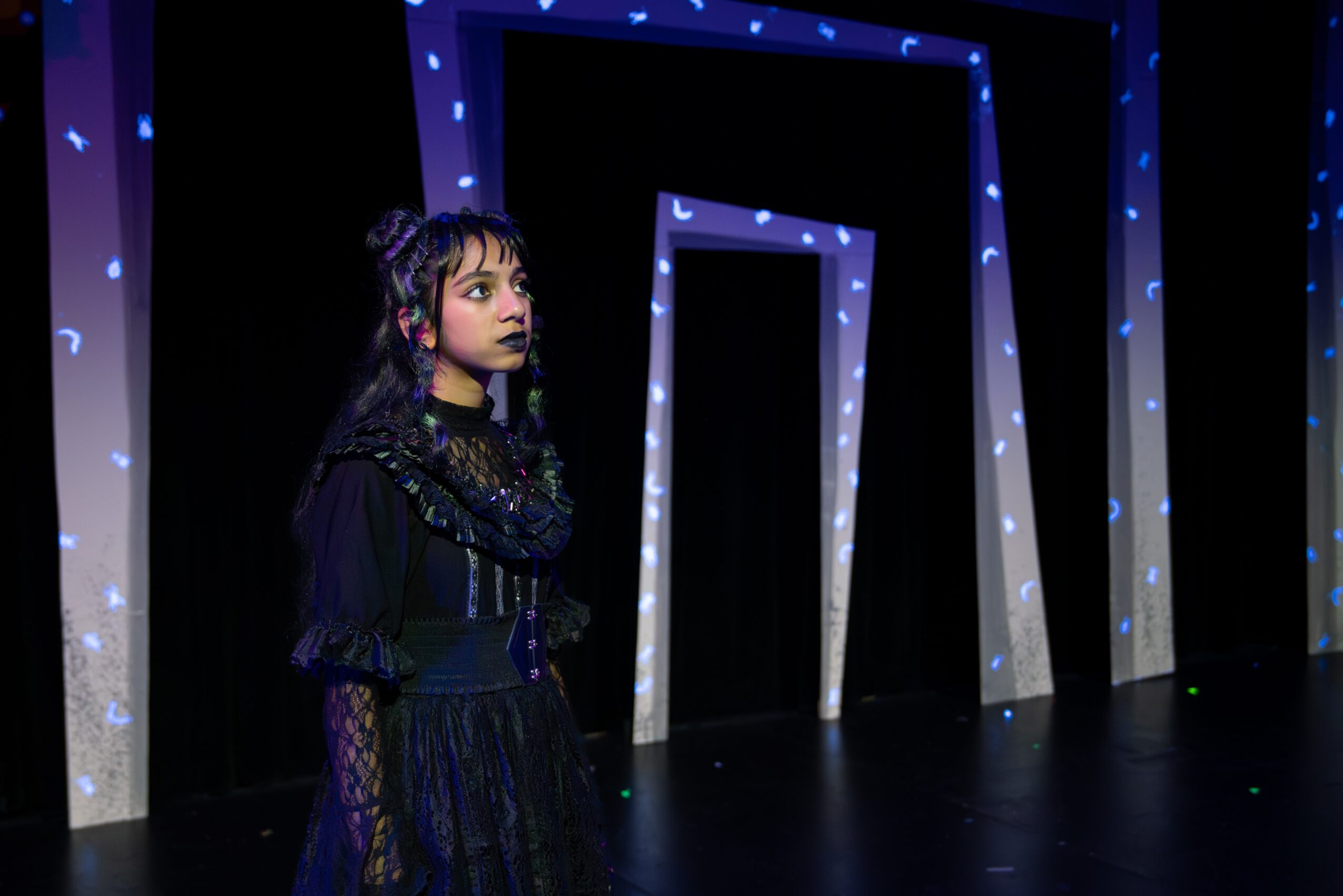 Young woman with dark hair and makeup stands in front of illuminated geometric frames on a dark stage.