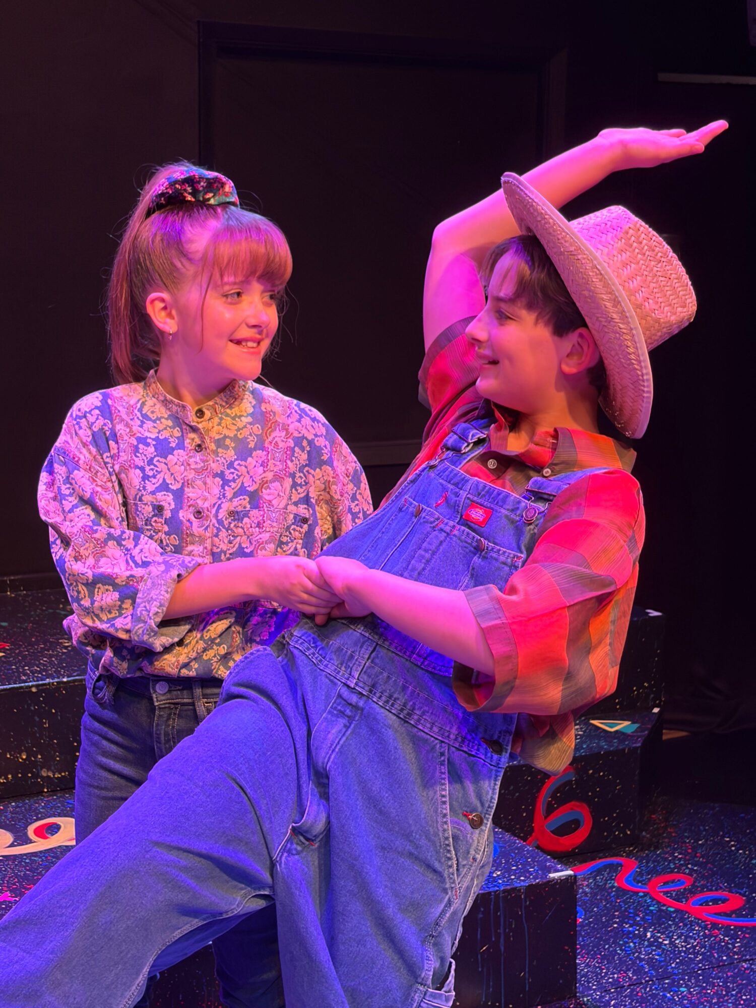Two children, one wearing a straw hat and plaid shirt, the other in a floral dress, are on stage with a dark background.