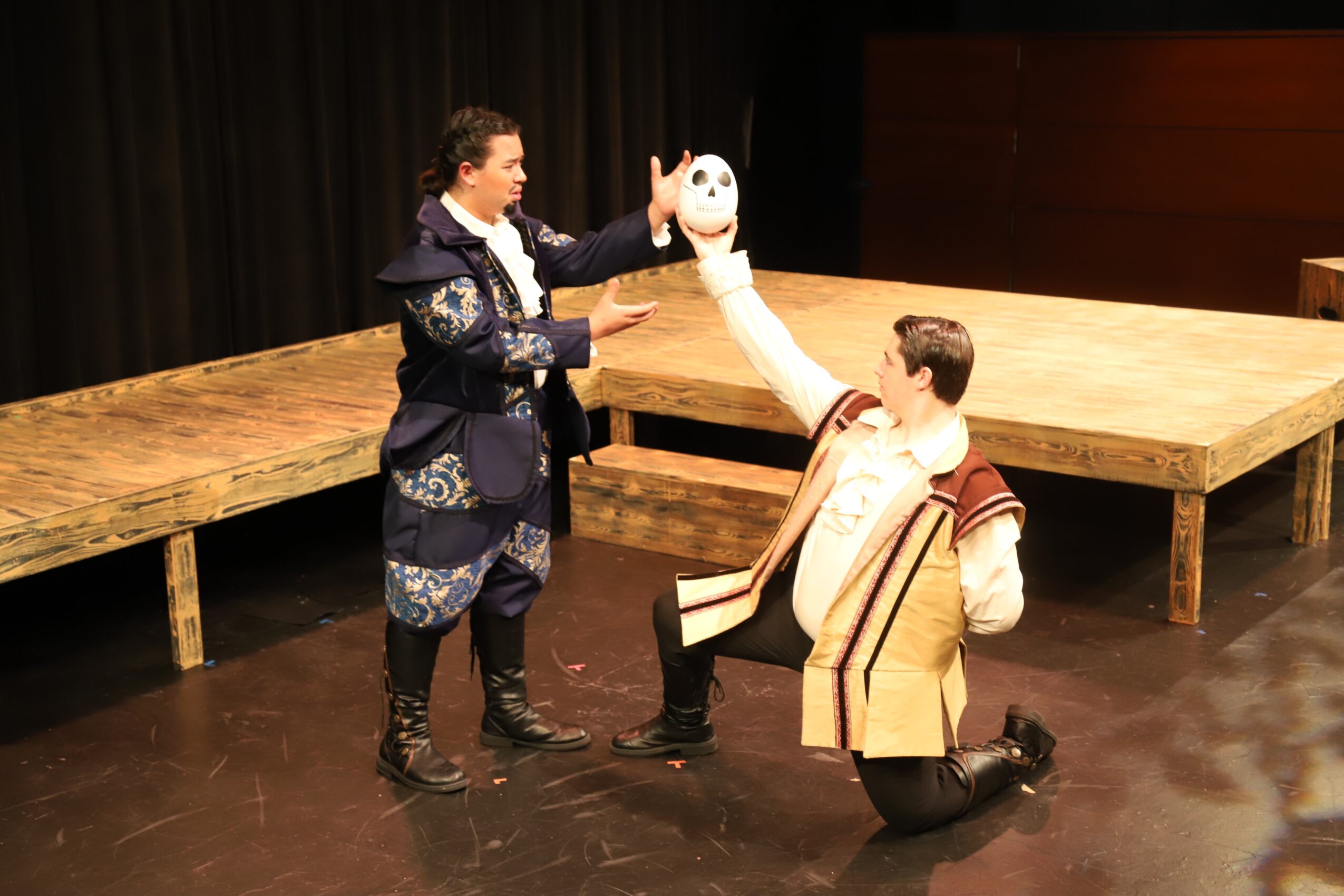 Two actors perform on stage, one with a puppet head, the other kneeling, with a wooden platform in the background.