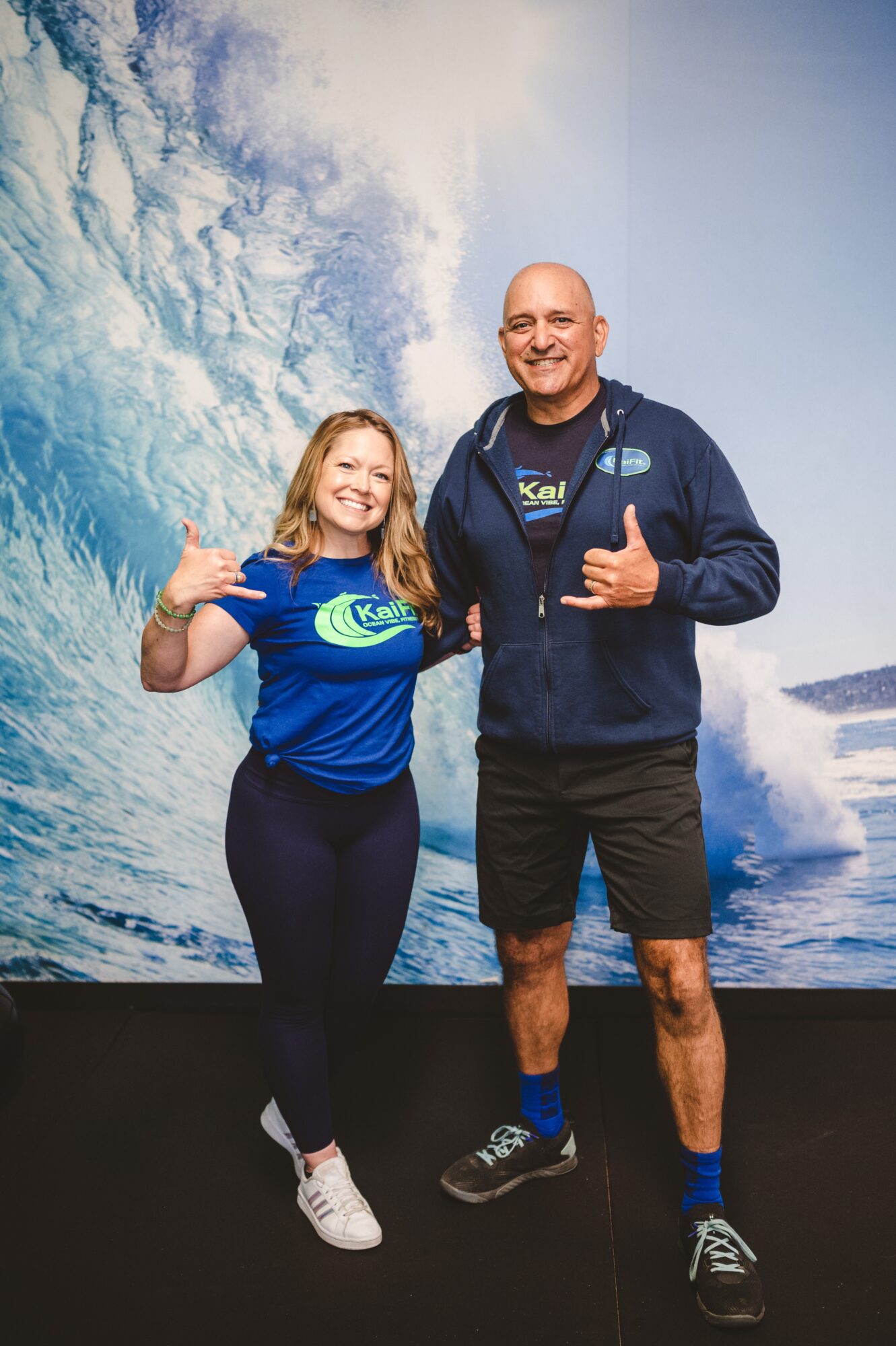 A smiling girl and man stand in front of a wave backdrop, both giving thumbs-up gestures, wearing blue shirts and casual shorts.