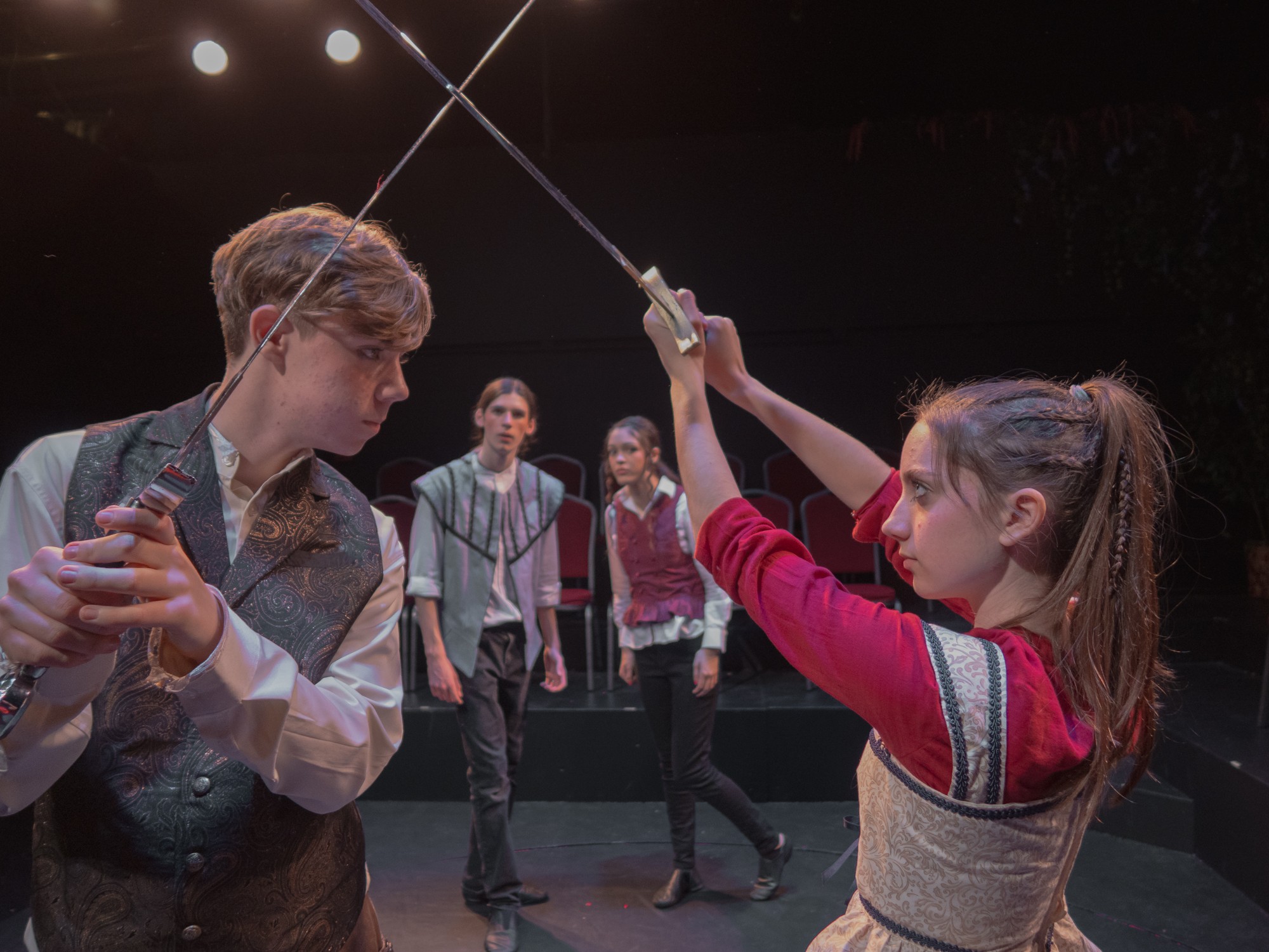 Girl in red and gray fencing outfit with a sword, aiming at a boy in a vest, with three people watching in background.