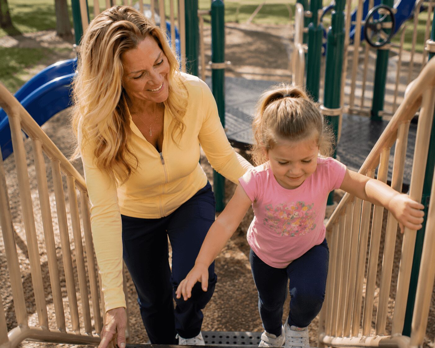 Woman and girl climbing playground stairs outdoors, smiling, surrounded by playground equipment.