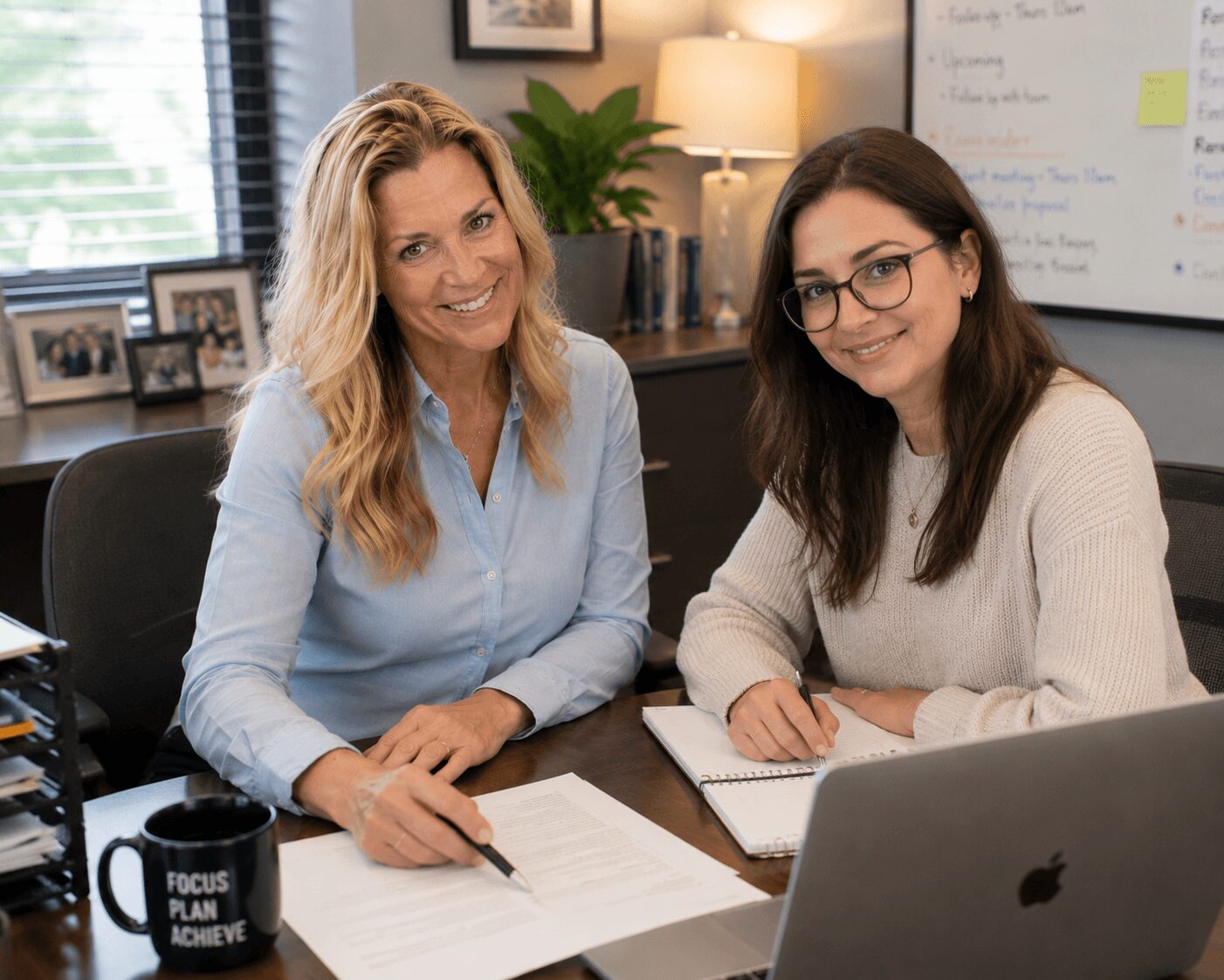 Two women sitting at a desk with notebooks, smiling, in an office with a whiteboard and window behind them.