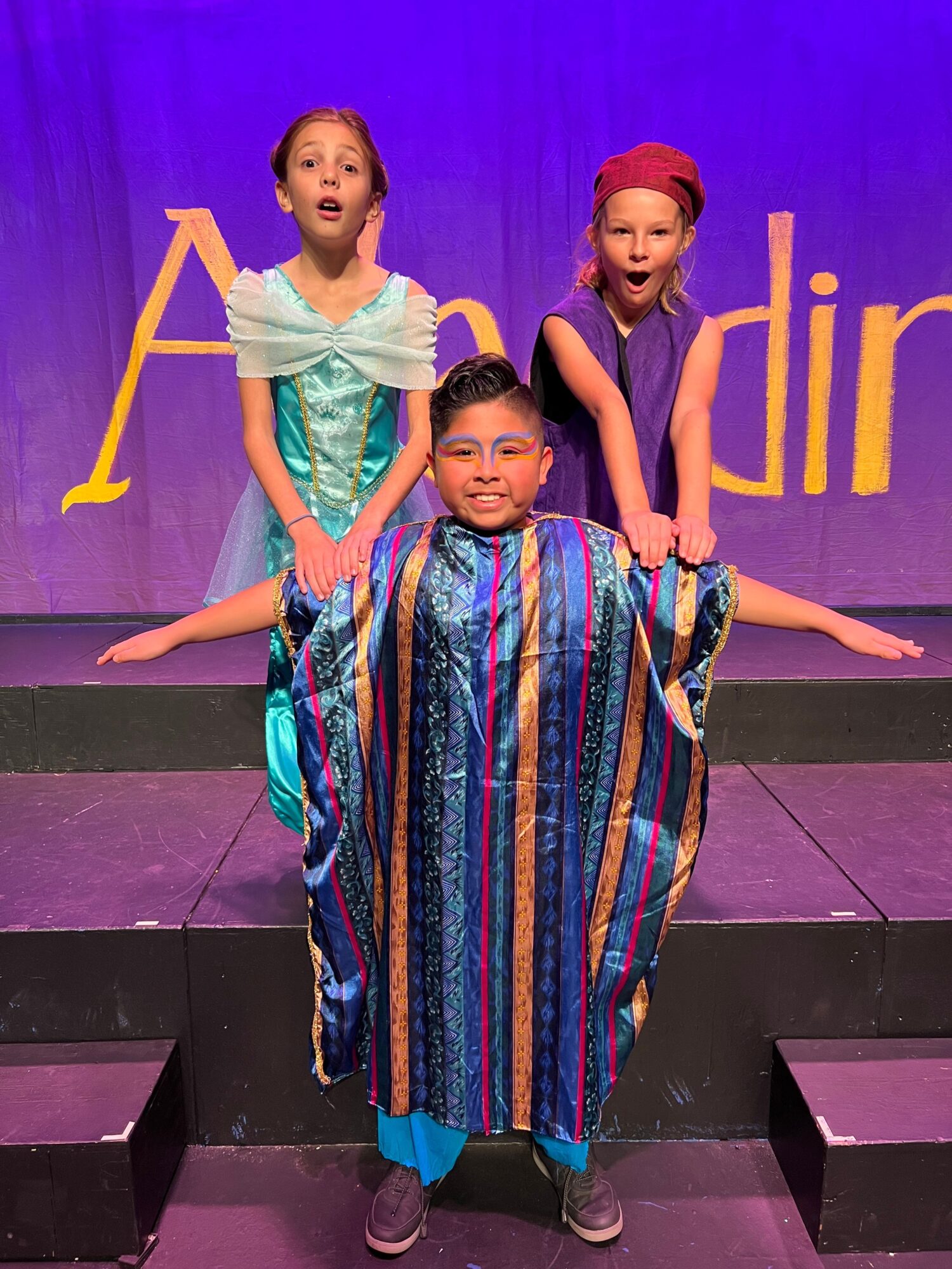 Three people on stage, two women and one girl, with purple and yellow background, posing with arms extended.