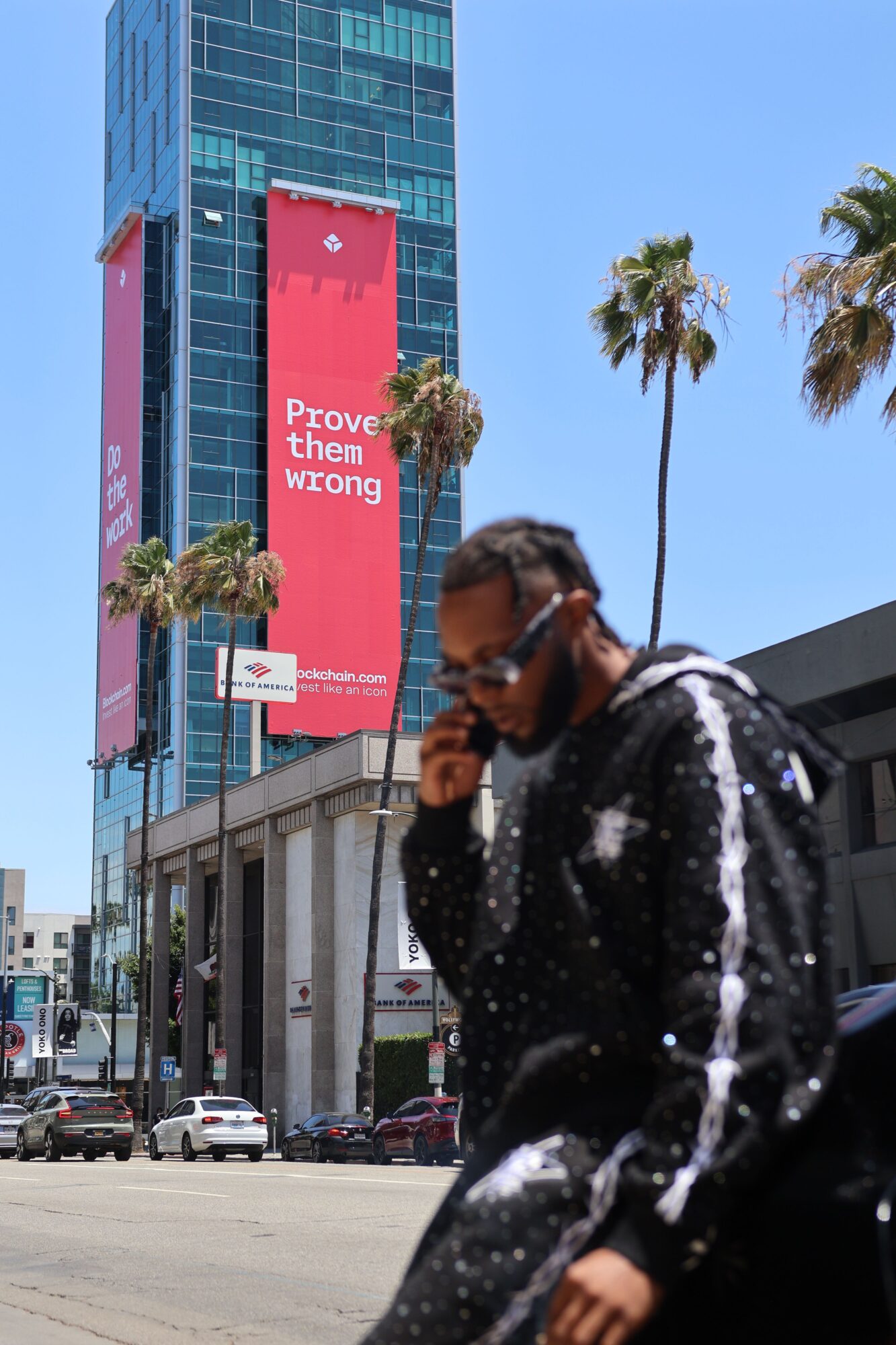 Person with dark hair and glasses wearing a black jacket, standing outdoors near buildings and palm trees.