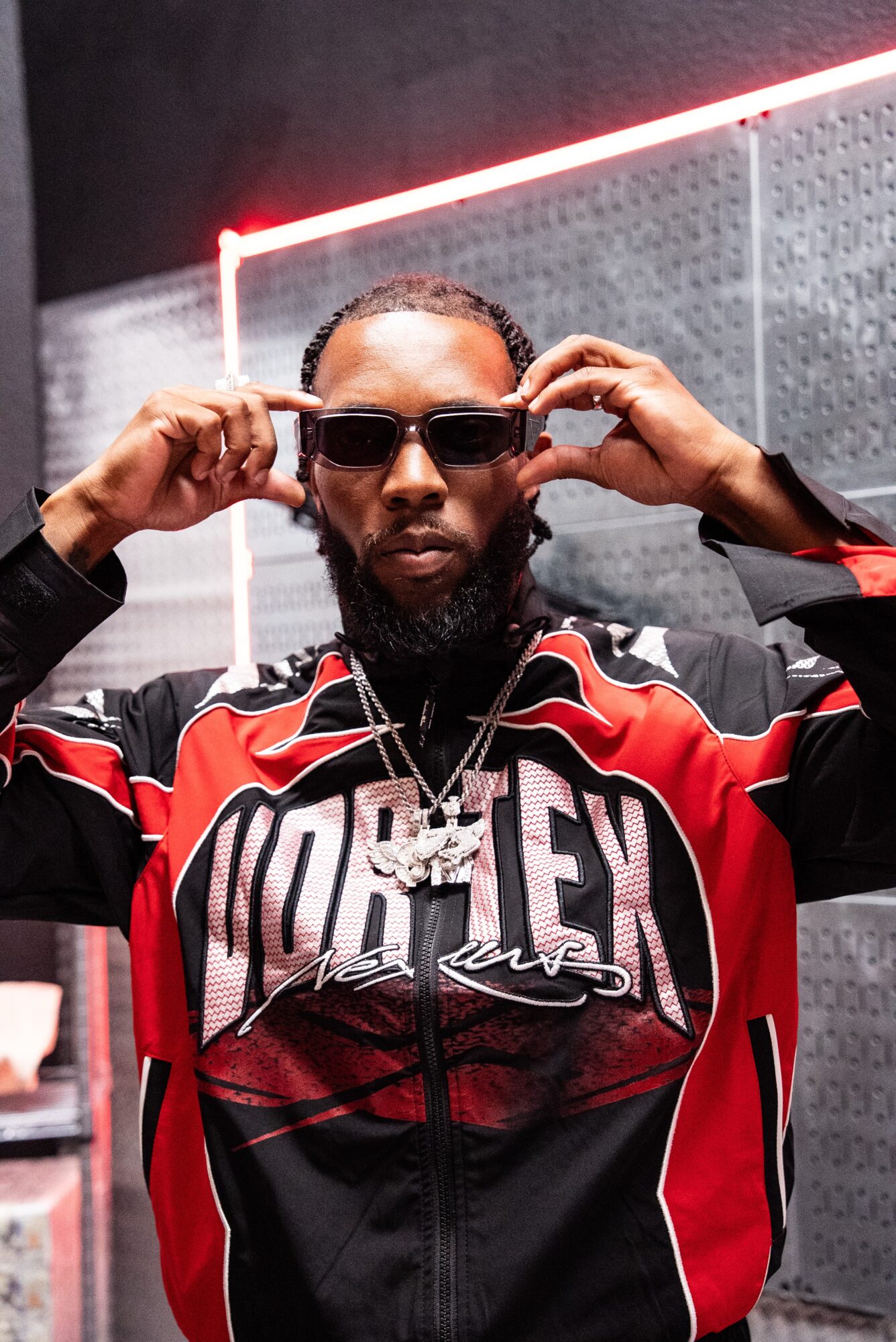 Person with sunglasses adjusting them, wearing a red and black jacket with a large logo, in a modern indoor setting.