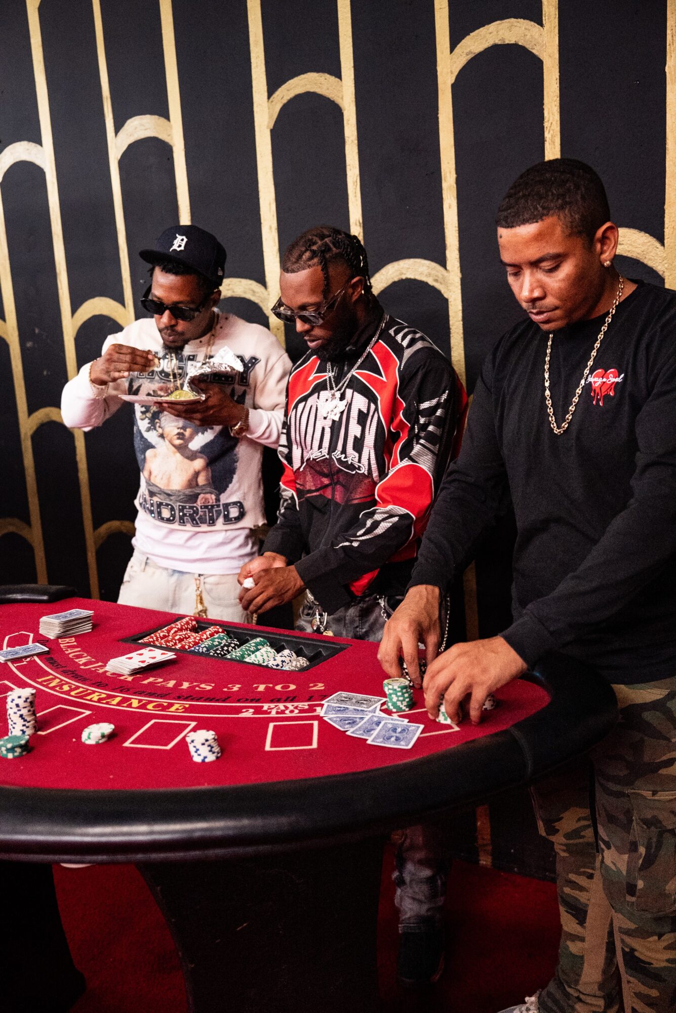 Three men stand around a red casino table with chips and cards, one is taking a photo, and two are playing.