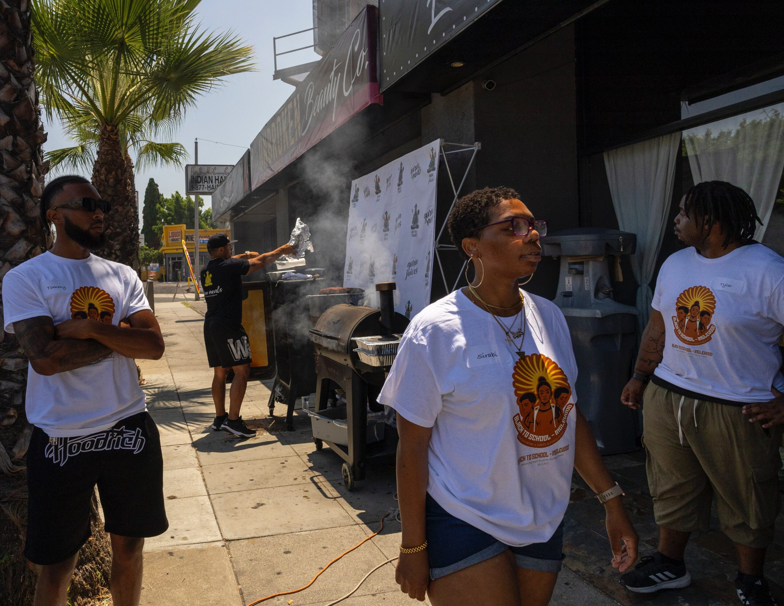 Group of people wearing white T-shirts with a logo, standing outside near a food stand with smoke, palm trees, and a building.