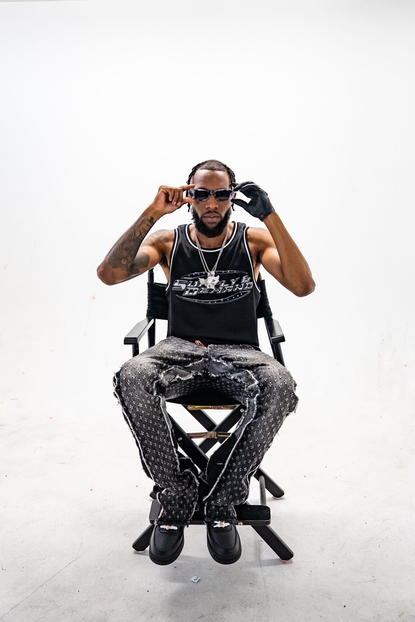Man sitting on a chair adjusting sunglasses, wearing patterned pants and a black tank top, against a plain white background.