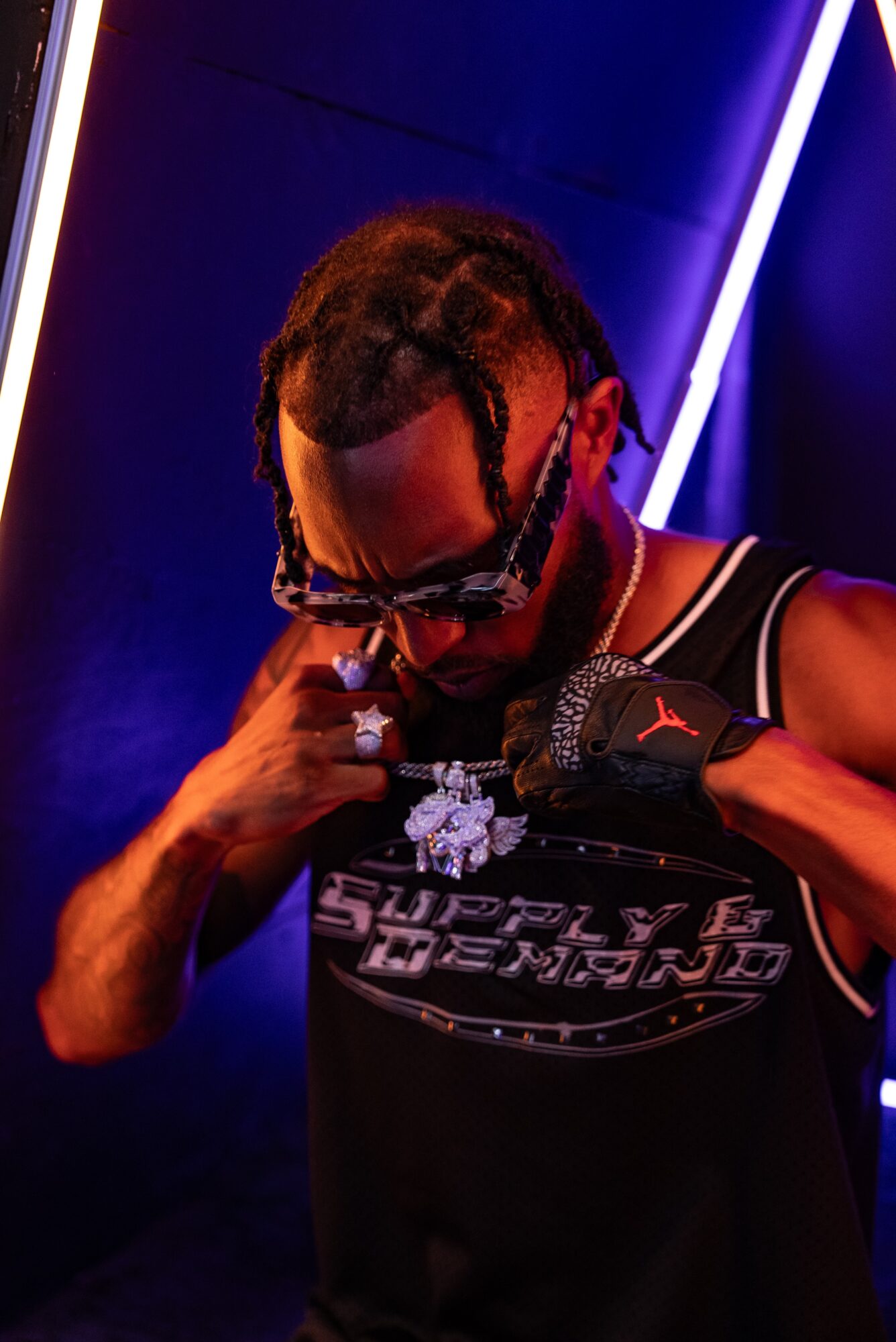 Person with braided hair, sunglasses, and a black sleeveless shirt adjusting a necklace, in a dark setting with neon lights.
