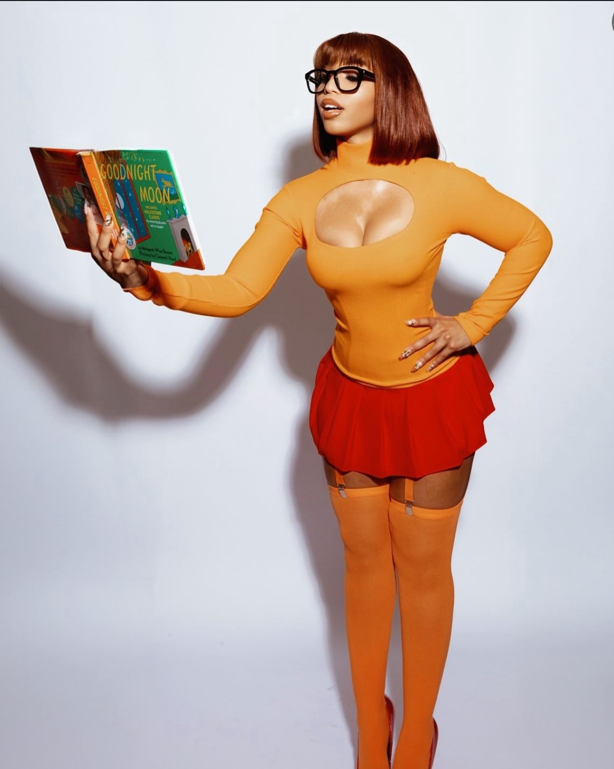 Woman with red hair, glasses, orange top, red skirt, and orange stockings holding a book, standing against a plain background.