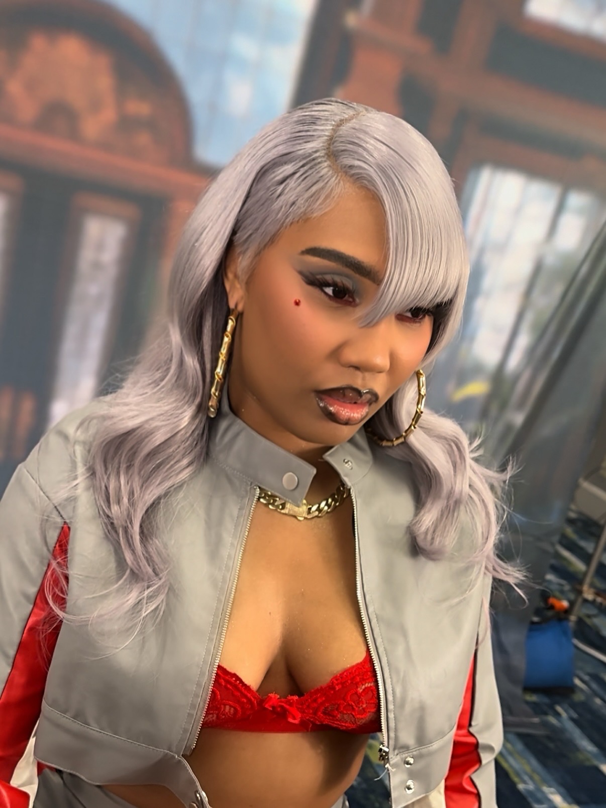 Woman with long, wavy silver hair, wearing large hoop earrings, a gray jacket with red sleeves, and a red lace top, indoors.