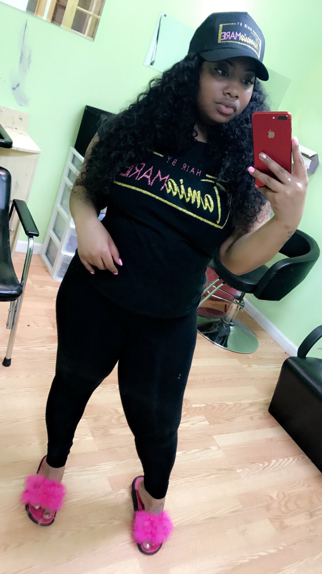 Young woman taking a mirror selfie in a room with green walls, wearing a black outfit, pink slippers, and a cap.