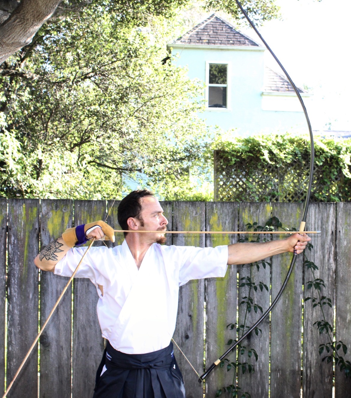Person practicing archery outdoors, drawing a bow, with a wooden fence and trees in background.