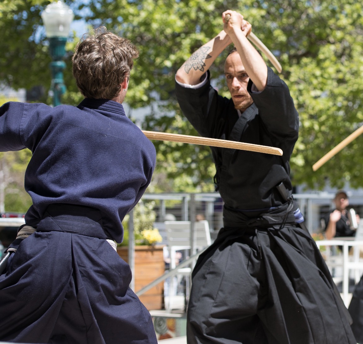 Two people practicing martial arts with wooden sticks outdoors, trees and a park setting in the background.