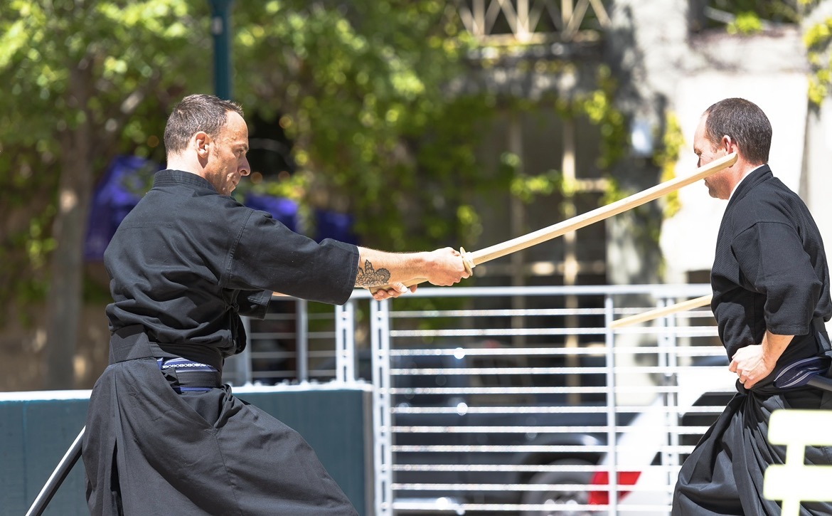 Two martial artists practicing with wooden sticks outdoors, one aiming at the other's face, trees and a fence in background.
