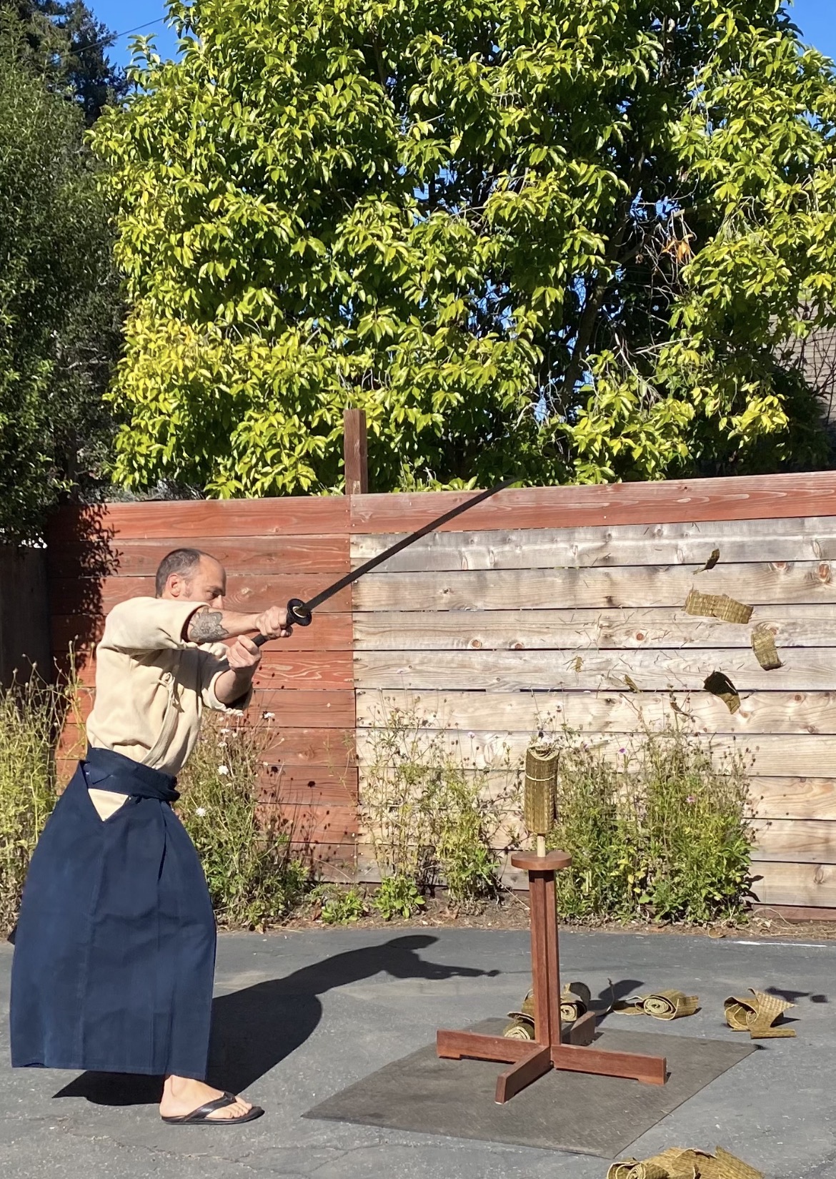 Person in traditional clothing practicing archery outdoors, aiming at a target with a bow and arrow, wooden fence and trees in background.