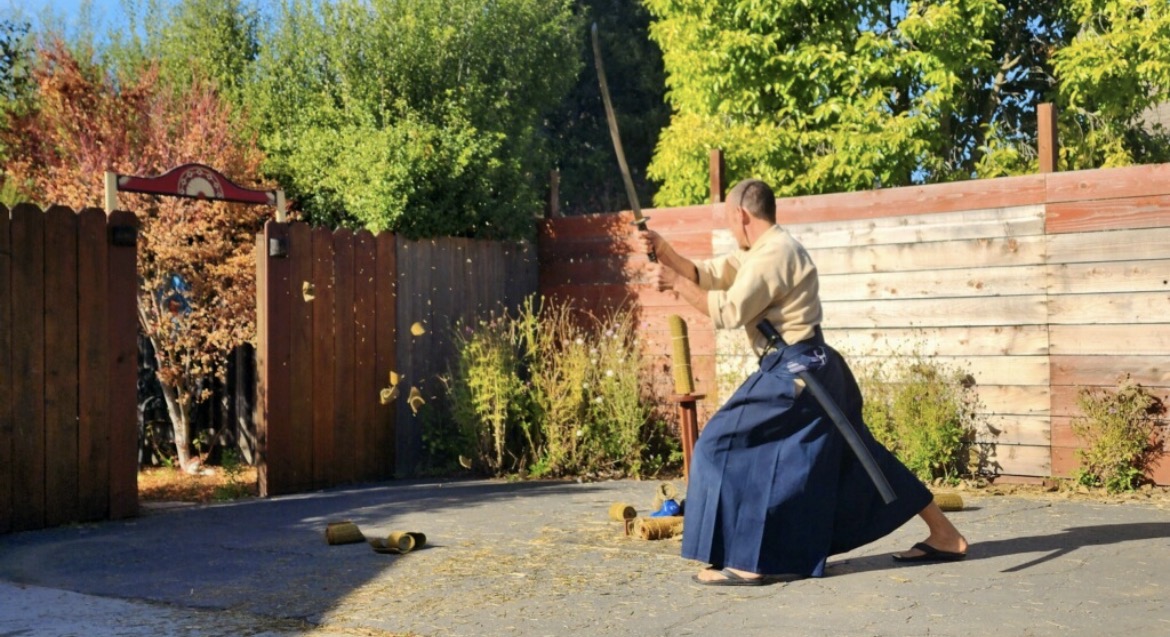 Person practicing martial arts outdoors in a yard with trees and wooden fences, wearing a traditional outfit, in sunlight.