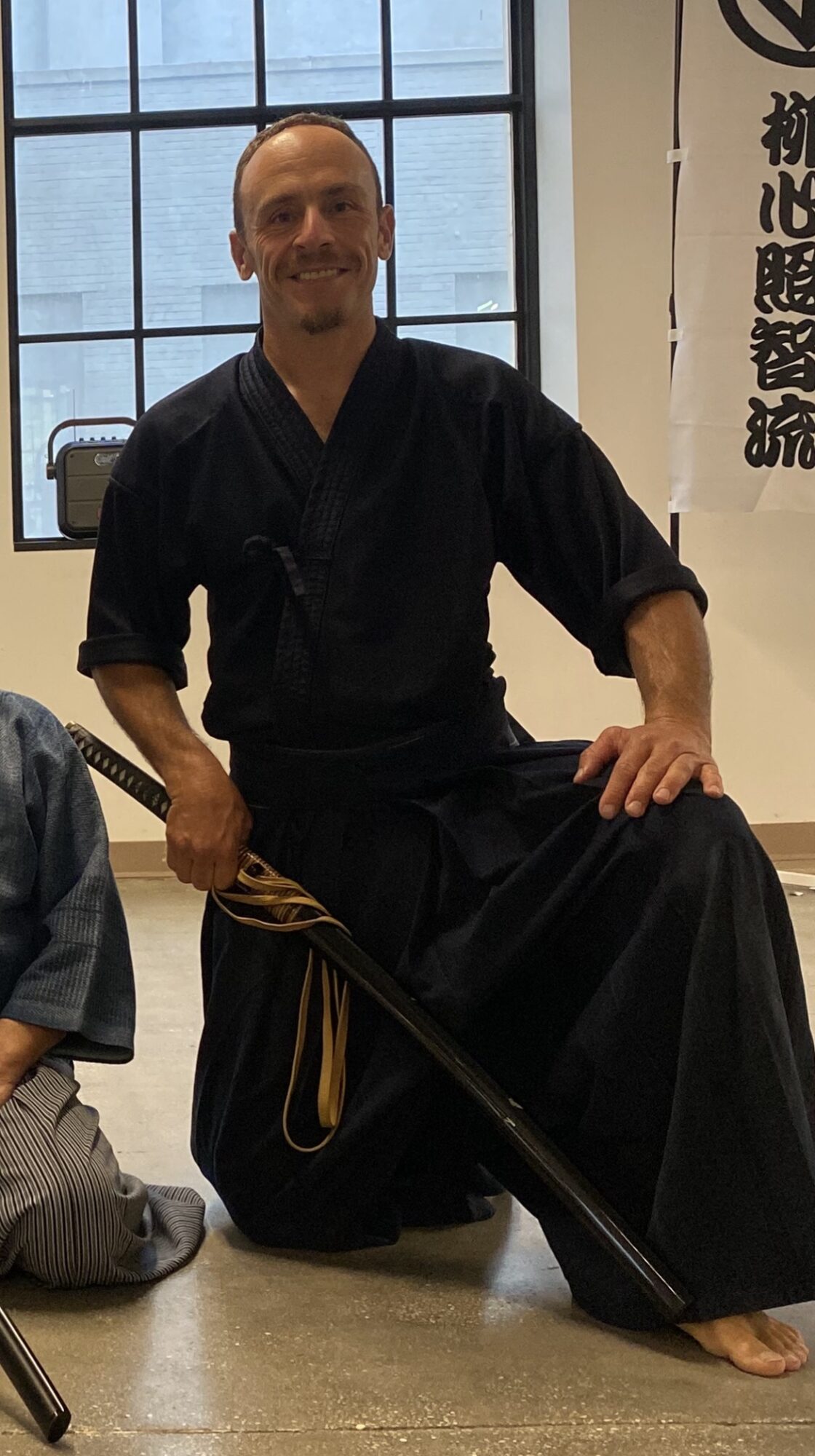 Person in traditional black martial arts attire kneeling with a sword, smiling, indoors near a window and a banner with Asian characters.
