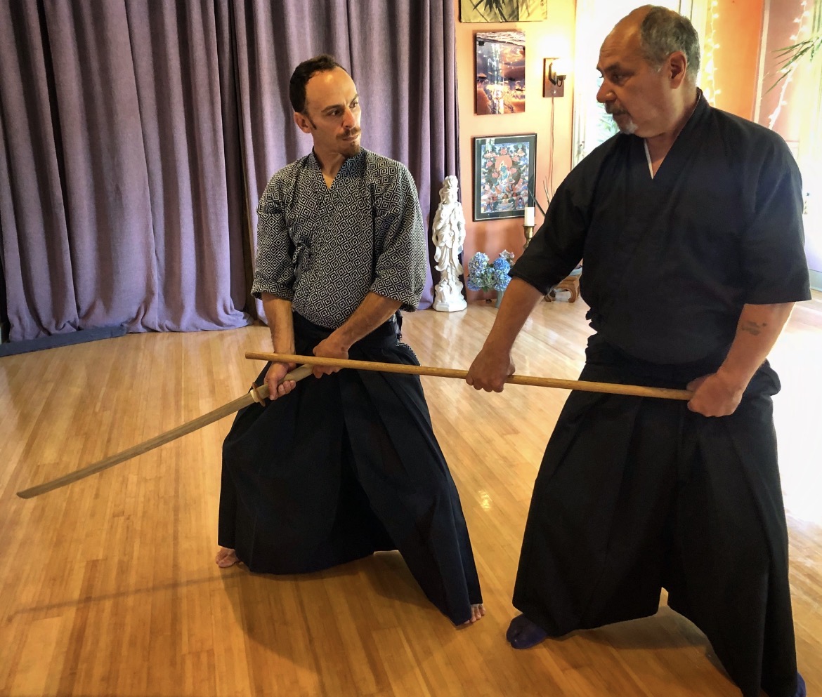 Two men practicing martial arts with a long staff in a room with wooden floor and purple curtains.
