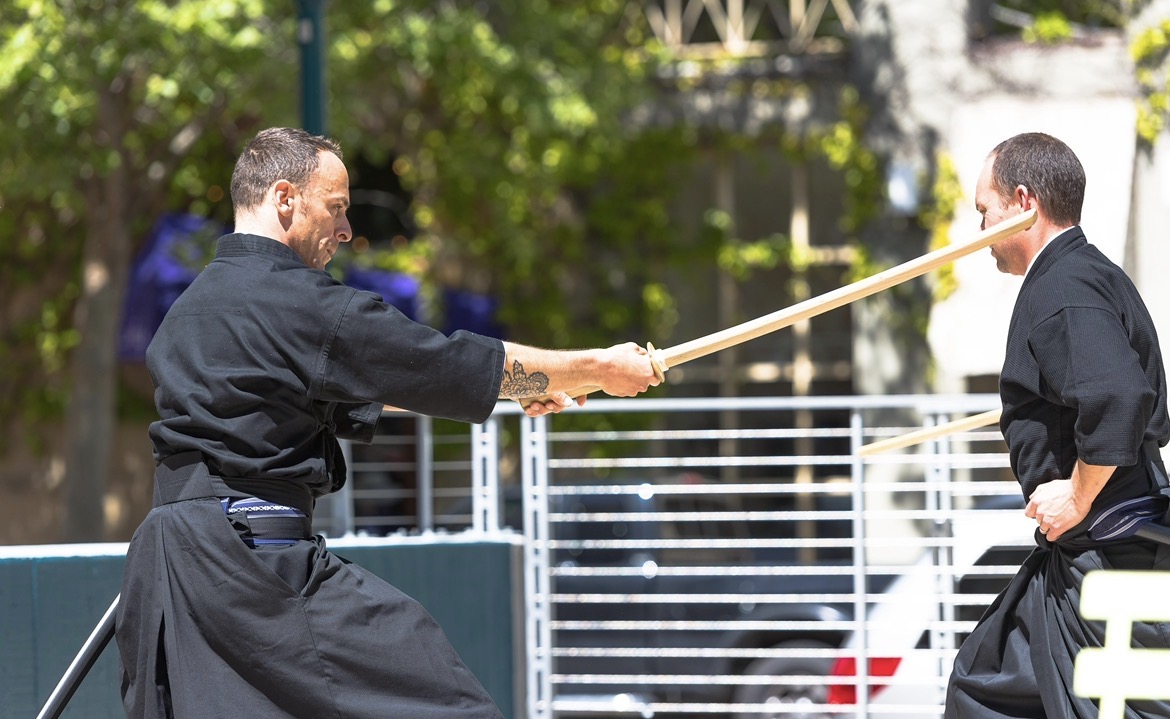 Two men in black traditional clothing engage in a martial arts demonstration outdoors, with one holding a wooden stick against the other's face.