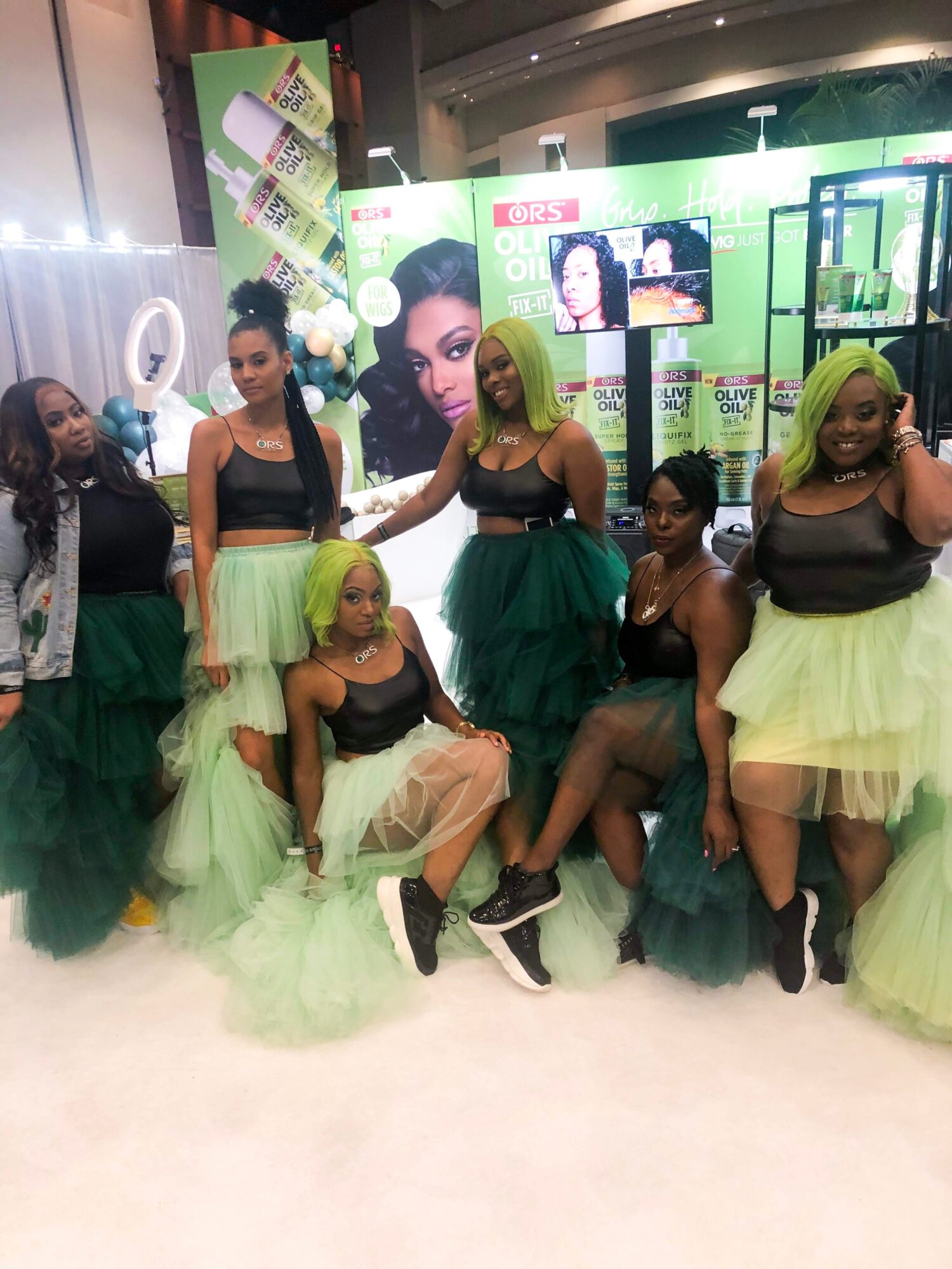 Group of six women in black and green outfits posing at an indoor event with promotional displays behind them.