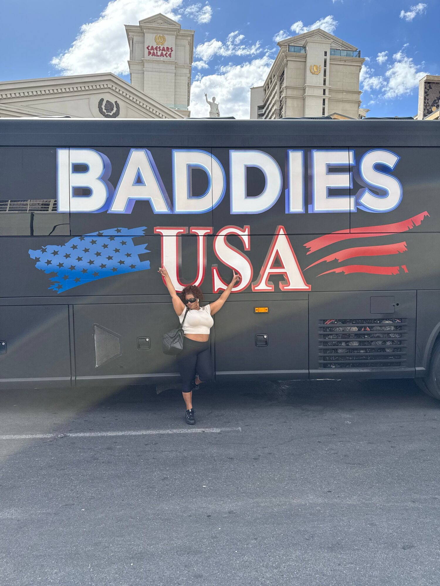Person standing in front of a bus with large text reading 'BADDIES USA' and a blue and red design, city buildings in background.