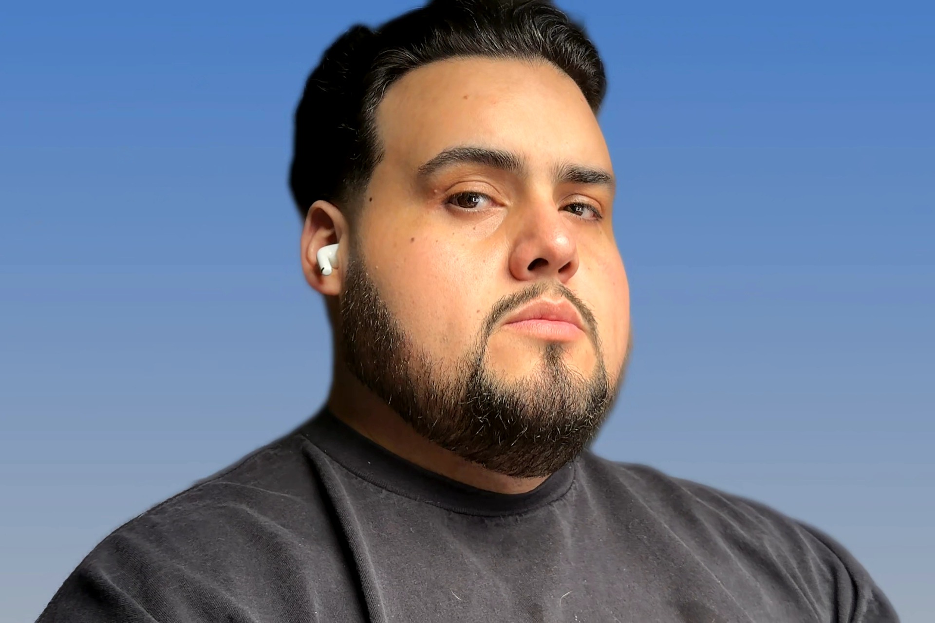 A man with dark hair, beard, and mustache, wearing a black shirt and white earbuds, looks serious against a blue background.