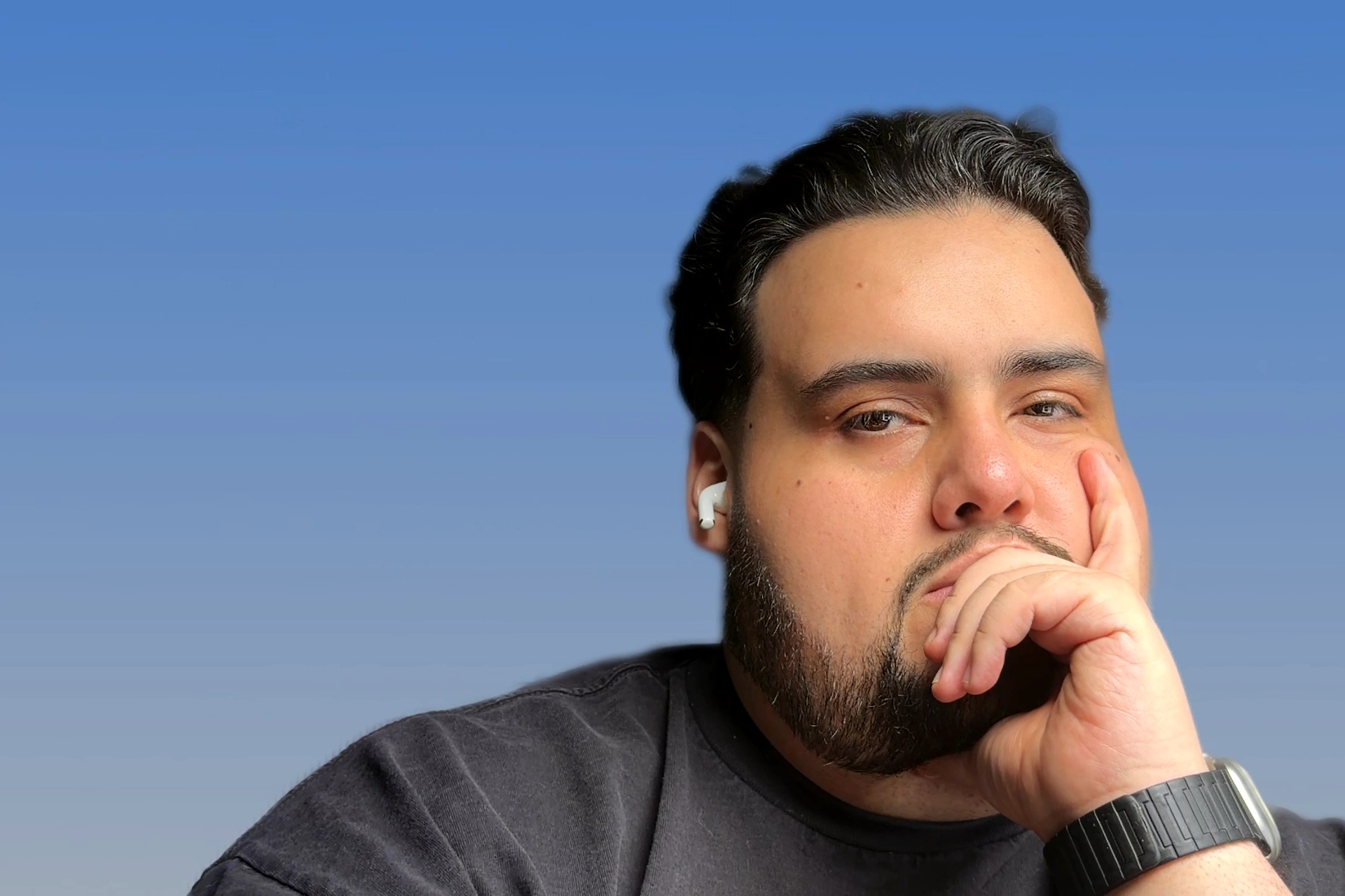 Man with dark hair and beard, wearing a black shirt, holding his hand near his face, against a blue background.