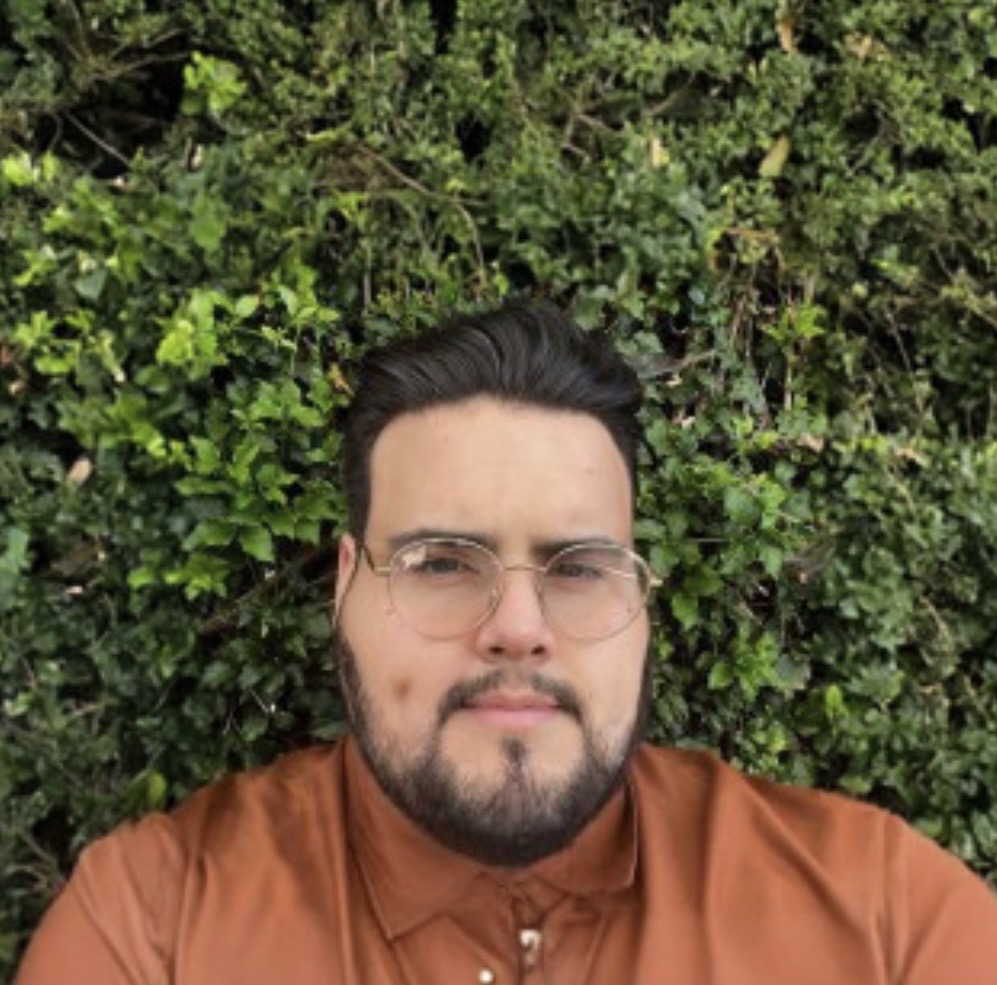 Man with glasses and beard lying on green leafy background, wearing a brown shirt.