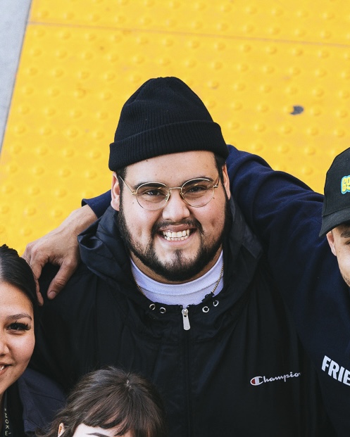 Smiling man wearing glasses, black beanie, and black jacket with a yellow background.