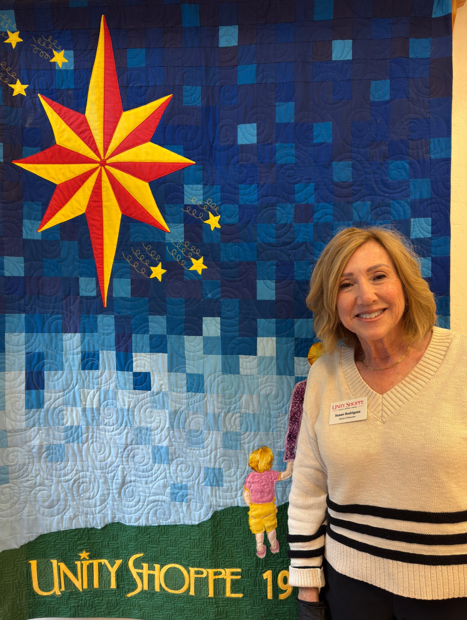 A woman with shoulder-length blonde hair smiling in front of a colorful quilt with a star and text, wearing a cream sweater with black stripes.