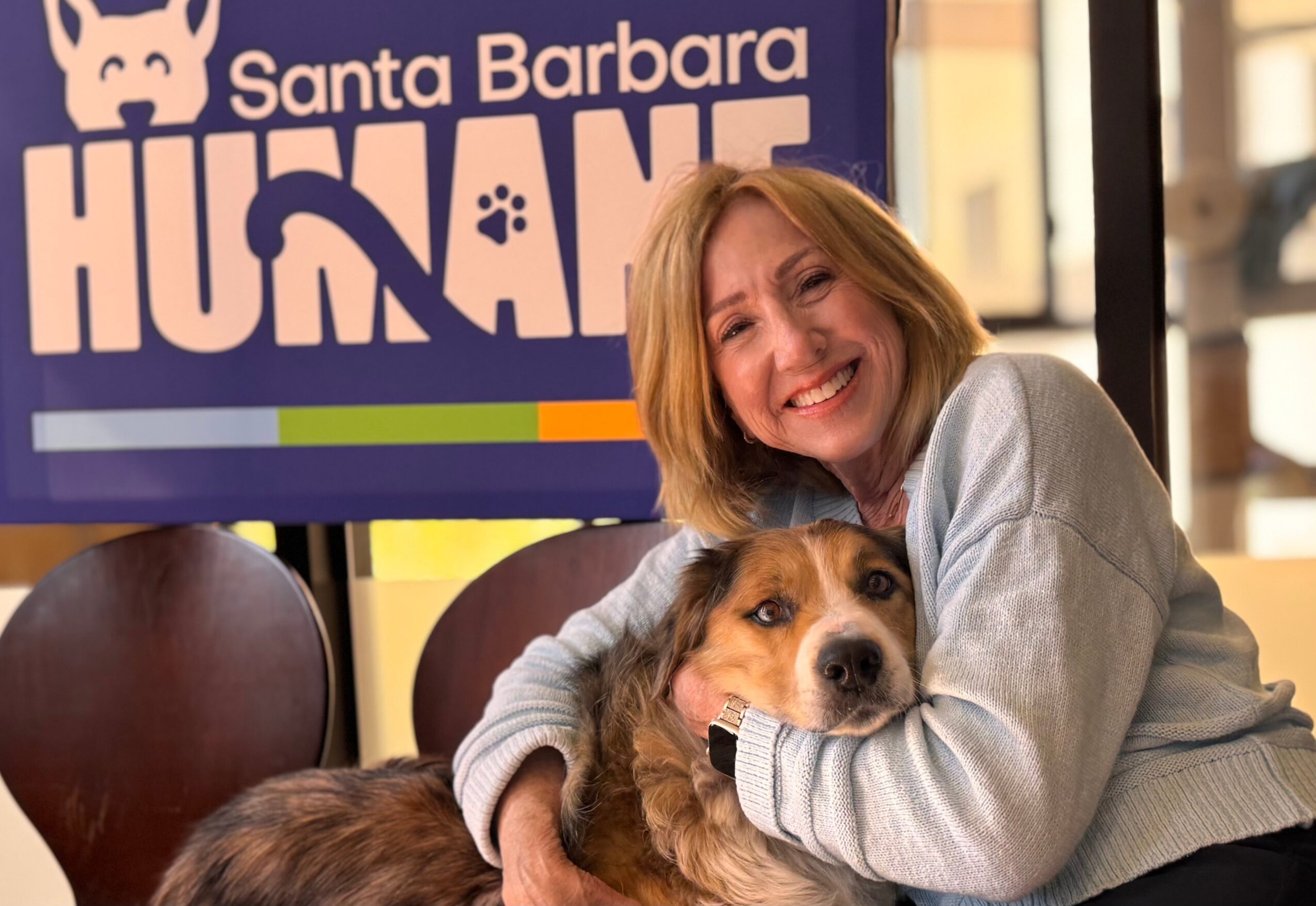 Woman hugging a dog in front of a Santa Barbara Hound sign, smiling. The dog is lying on a table.