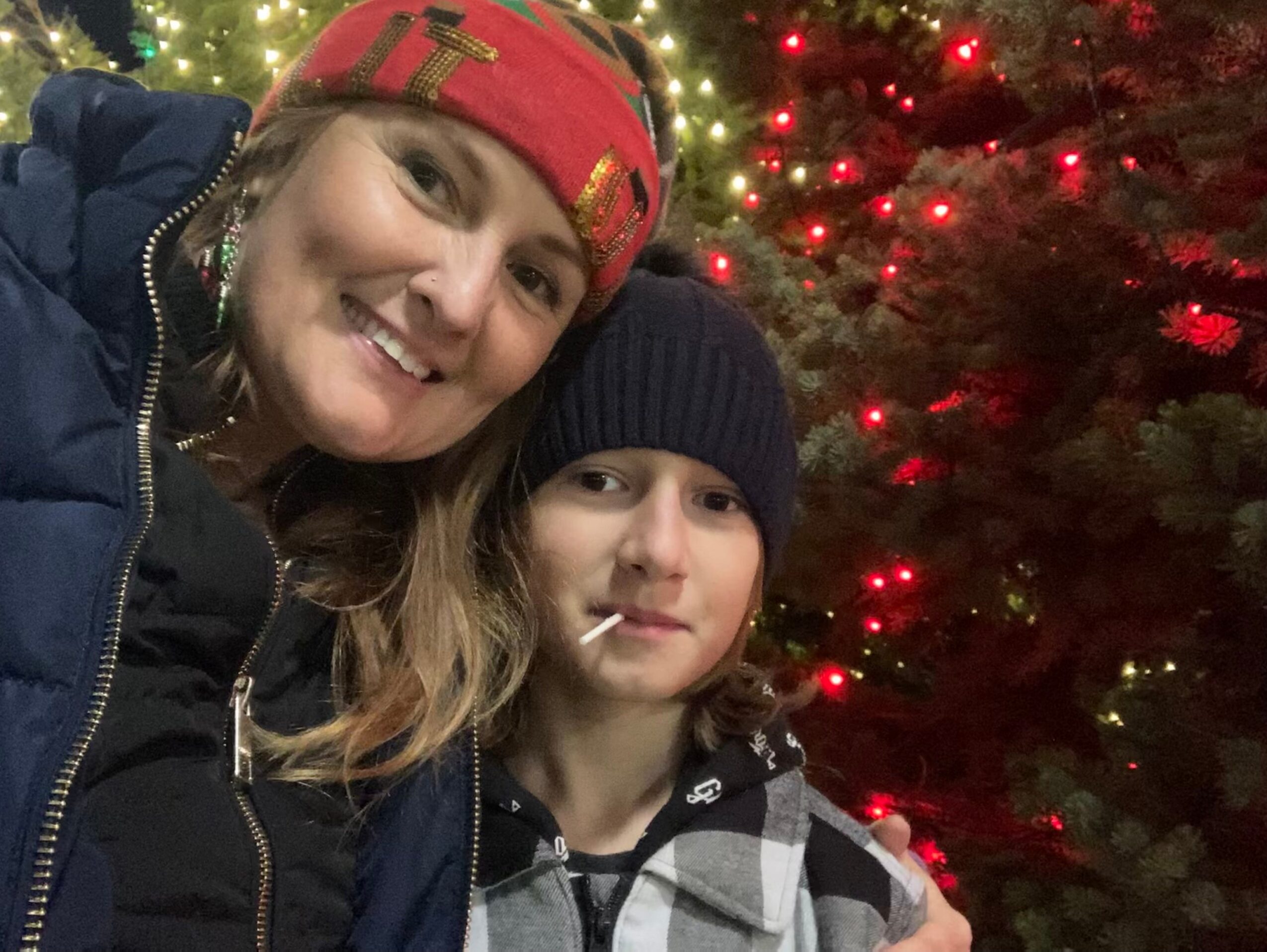 Two people smiling in front of a decorated Christmas tree with red and gold lights.