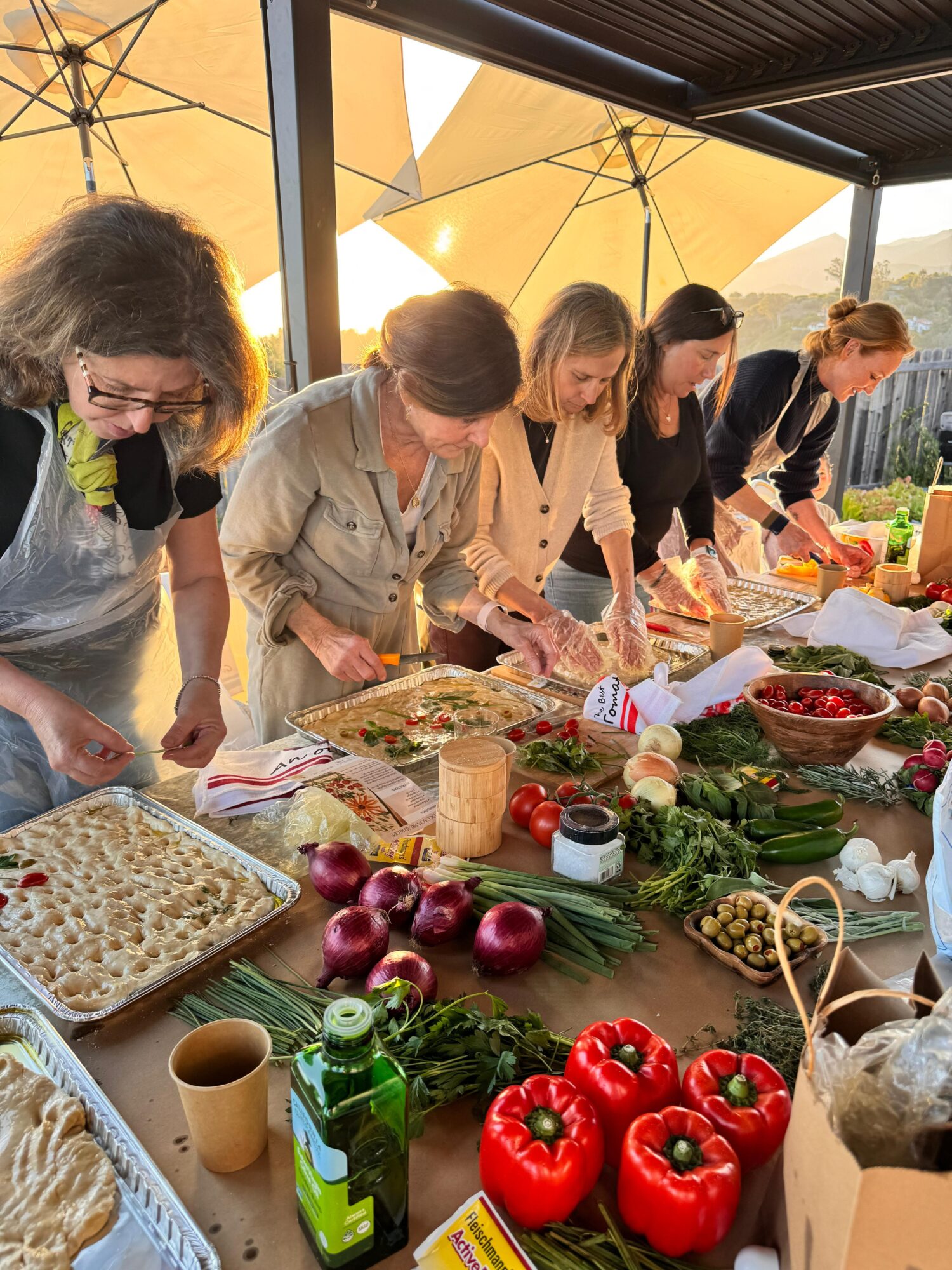 Group of women preparing food at a table outdoors with vegetables and ingredients, under a large yellow umbrella.
