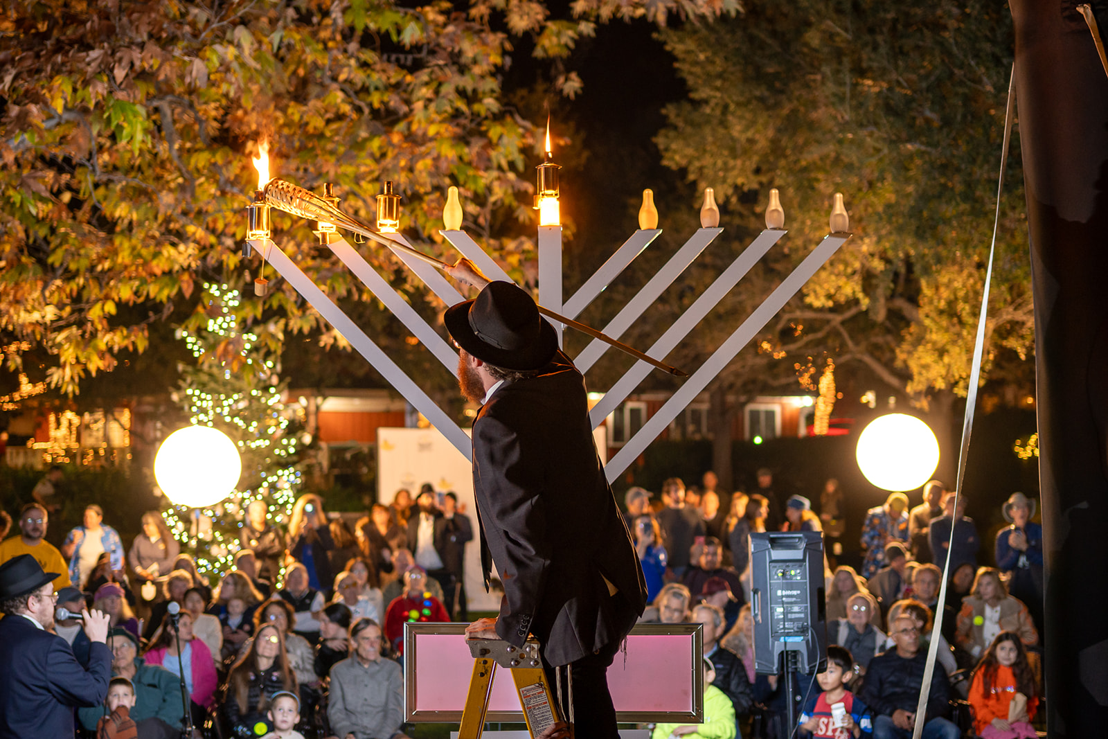 Person wearing a hat stands in front of a large menorah during a celebration at night, with a crowd and festive lights.