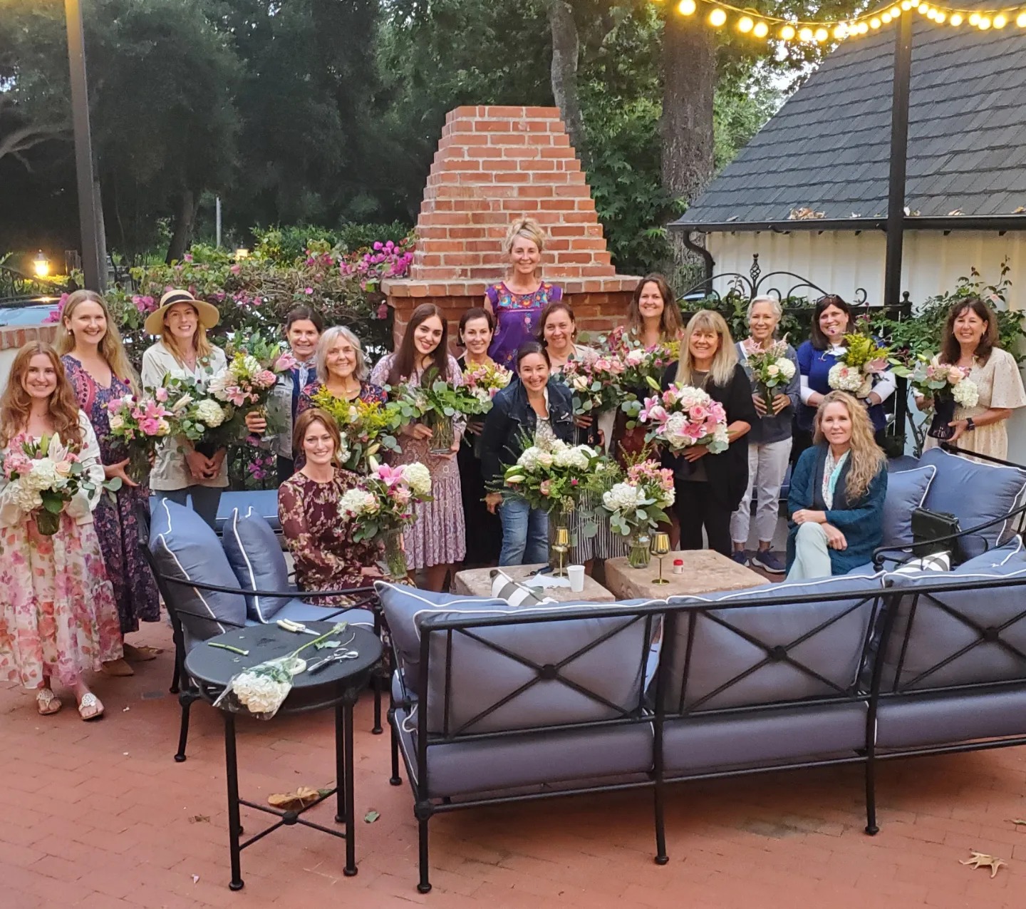 Group of women gathered outdoors with flowers, sitting and standing around a table, in front of a brick chimney and house.