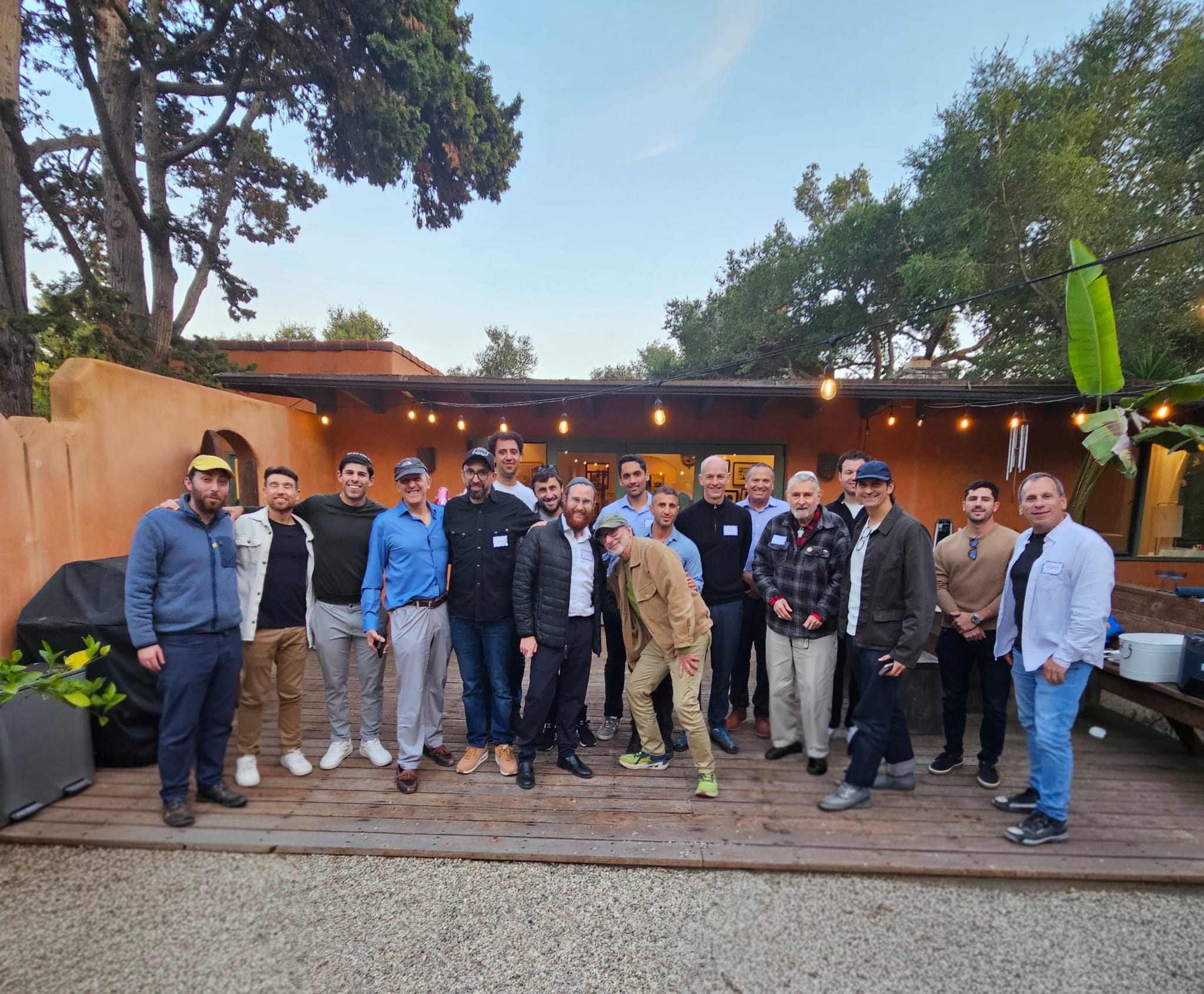 Group of people standing outdoors on a wooden deck with string lights and trees in the background.
